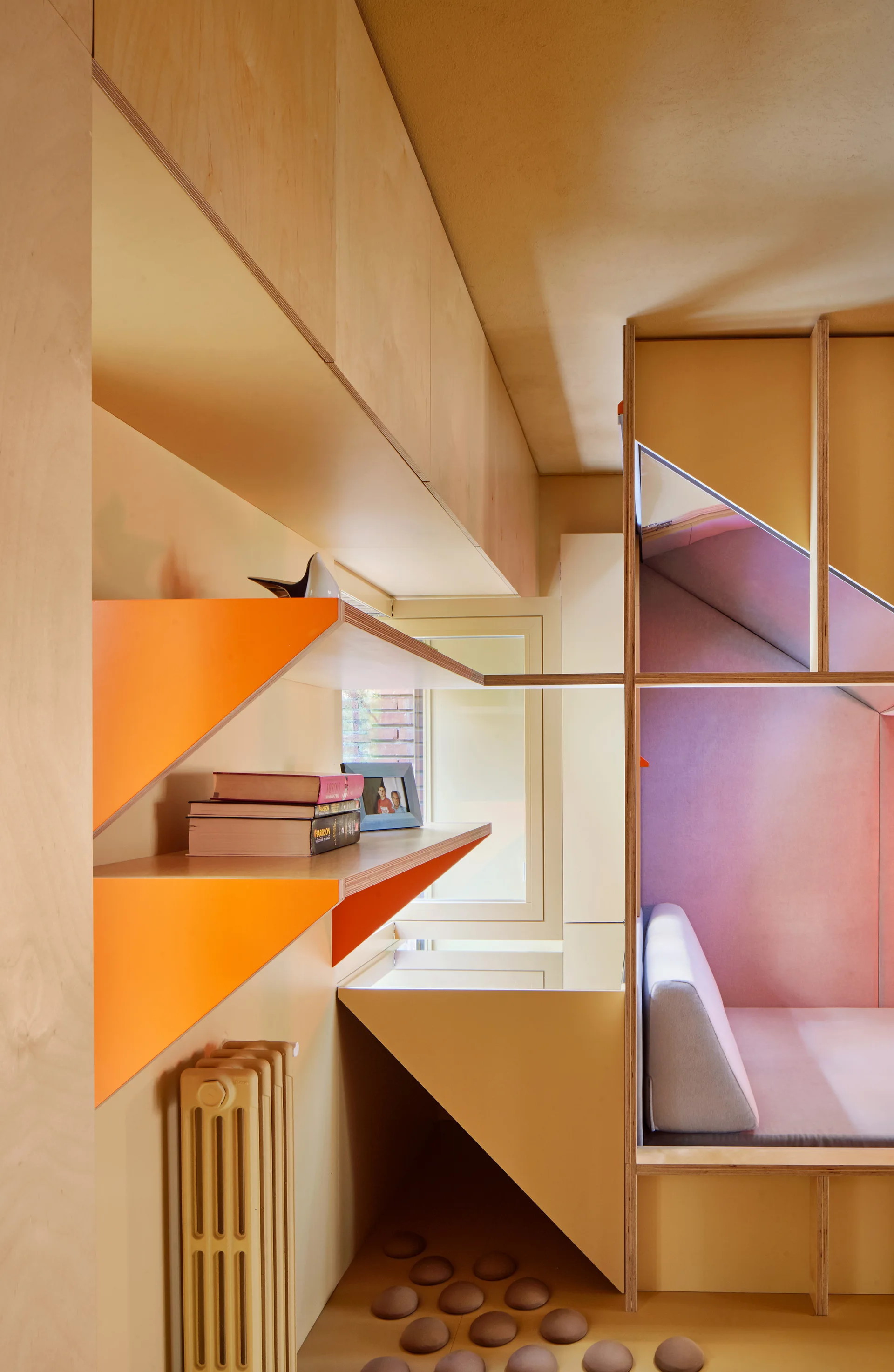 A custom light wood plywood built-in shelving unit featuring bright orange-painted shelf brackets and a cozy pink upholstered seating nook.