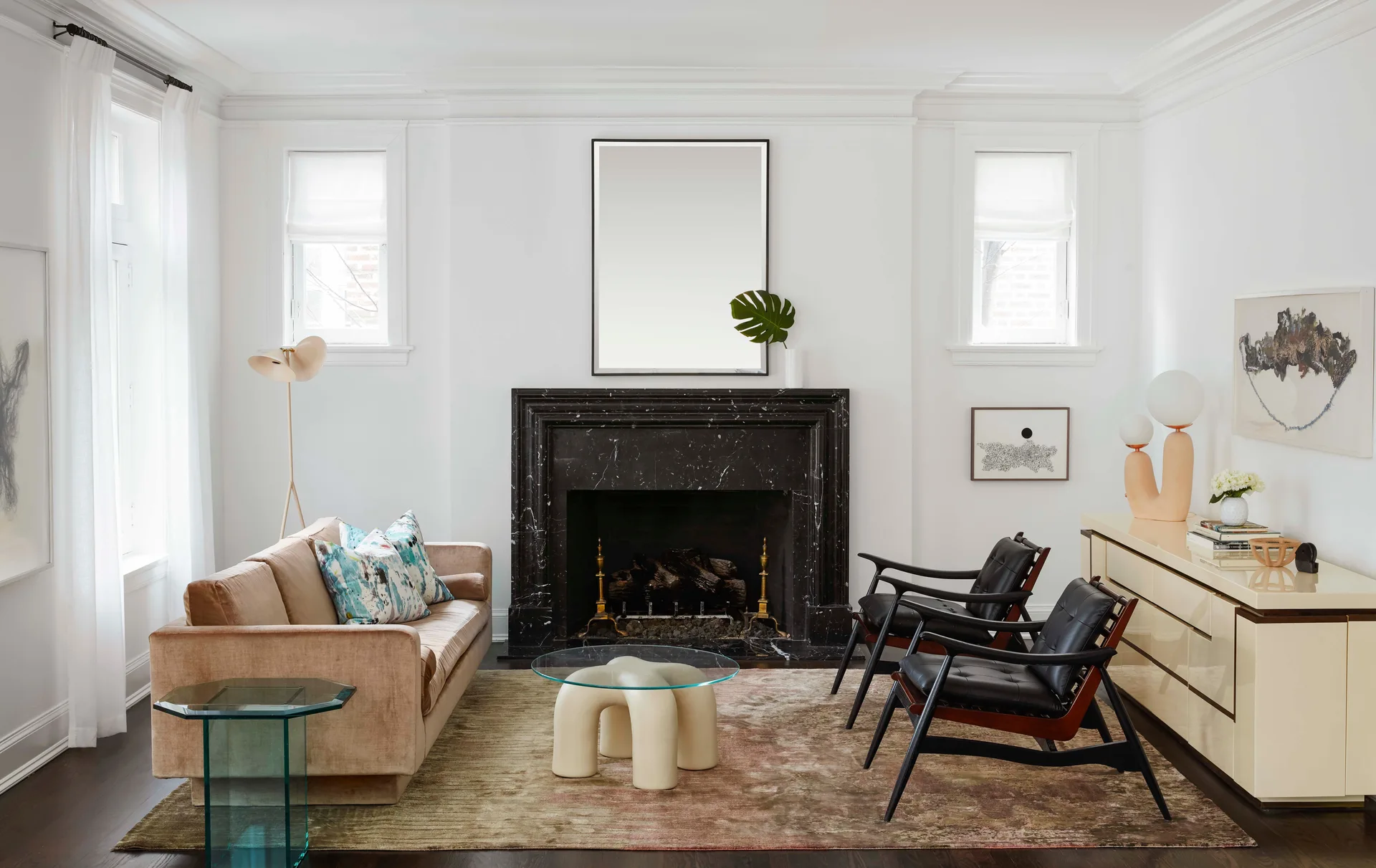 A white living room features a black marble fireplace flanked by windows, a velvet sofa, two leather armchairs, and a glass coffee table on an area rug.