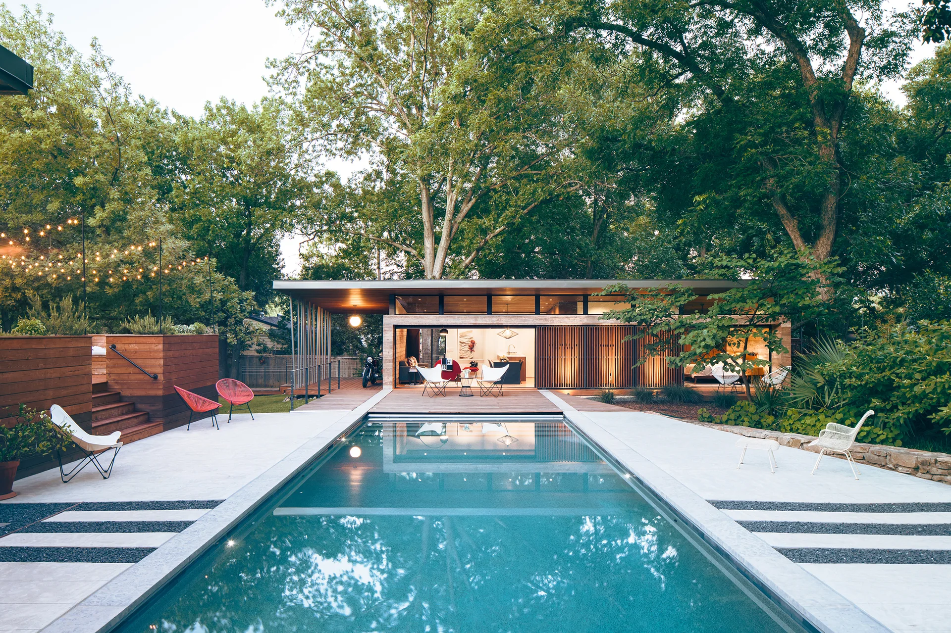 A rectangular swimming pool sits in front of a modern flat-roof house featuring large sliding glass walls, wood decking, and slatted screens.