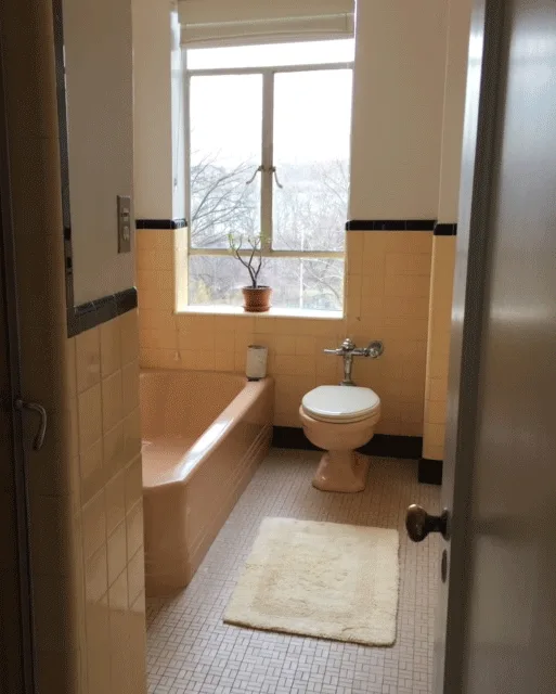 A vintage bathroom featuring pale yellow wall tiles with a thin black border trim, a built-in peach bathtub, a toilet, and a central window.