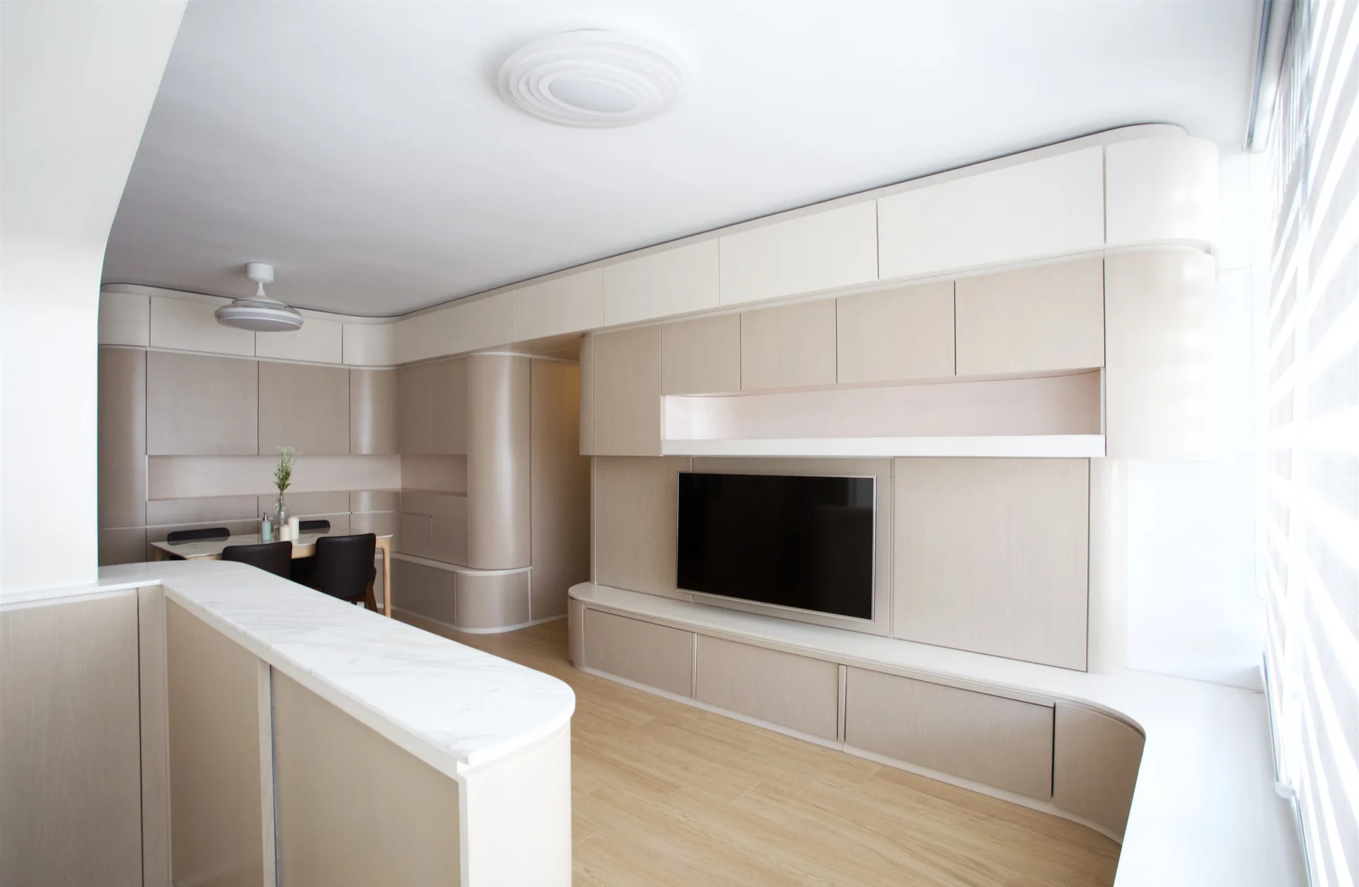 A modern living room features seamless light beige built-in cabinetry with rounded edges, a mounted television, and a marble-topped island.