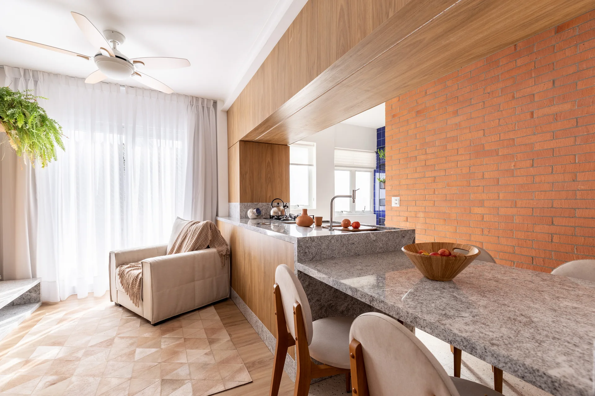 A bright kitchen features a stone-topped island with a wooden base, an exposed orange brick wall, and a beige armchair near a window with sheer drapes.