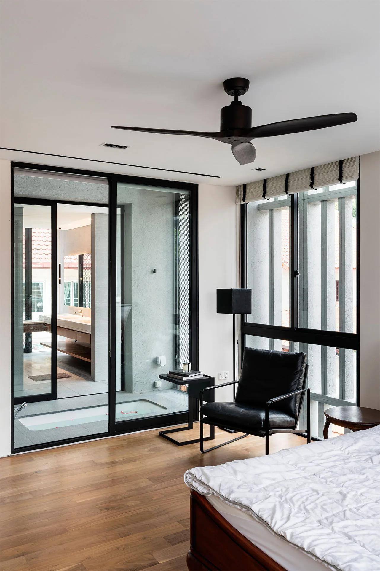 A minimalist bedroom features a black leather armchair, hardwood floors, and a glass sliding door looking into an ensuite bathroom.