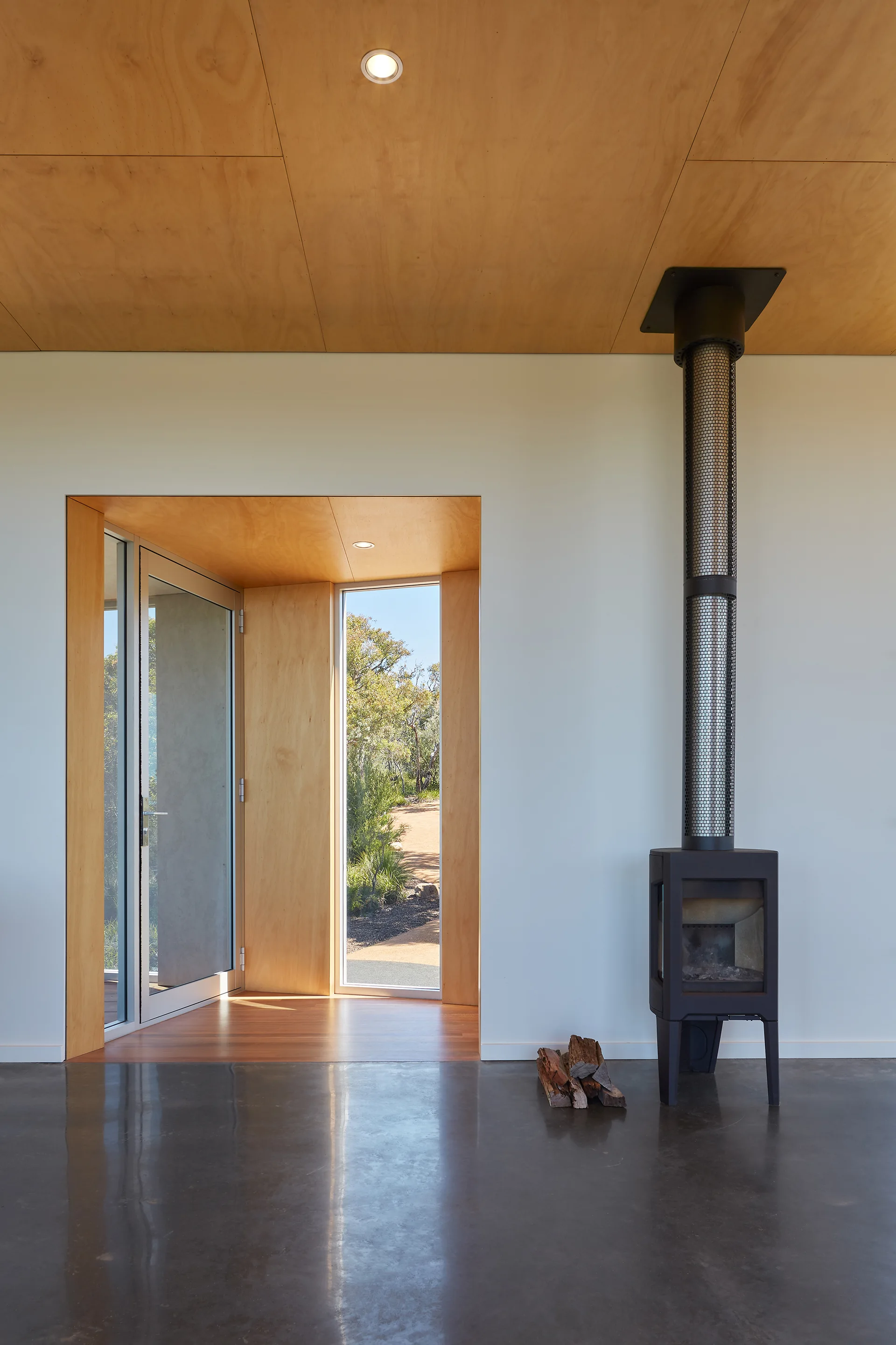 A minimalist interior features a black freestanding stove, a warm plywood ceiling, and polished concrete floors leading to a wood-framed entryway.