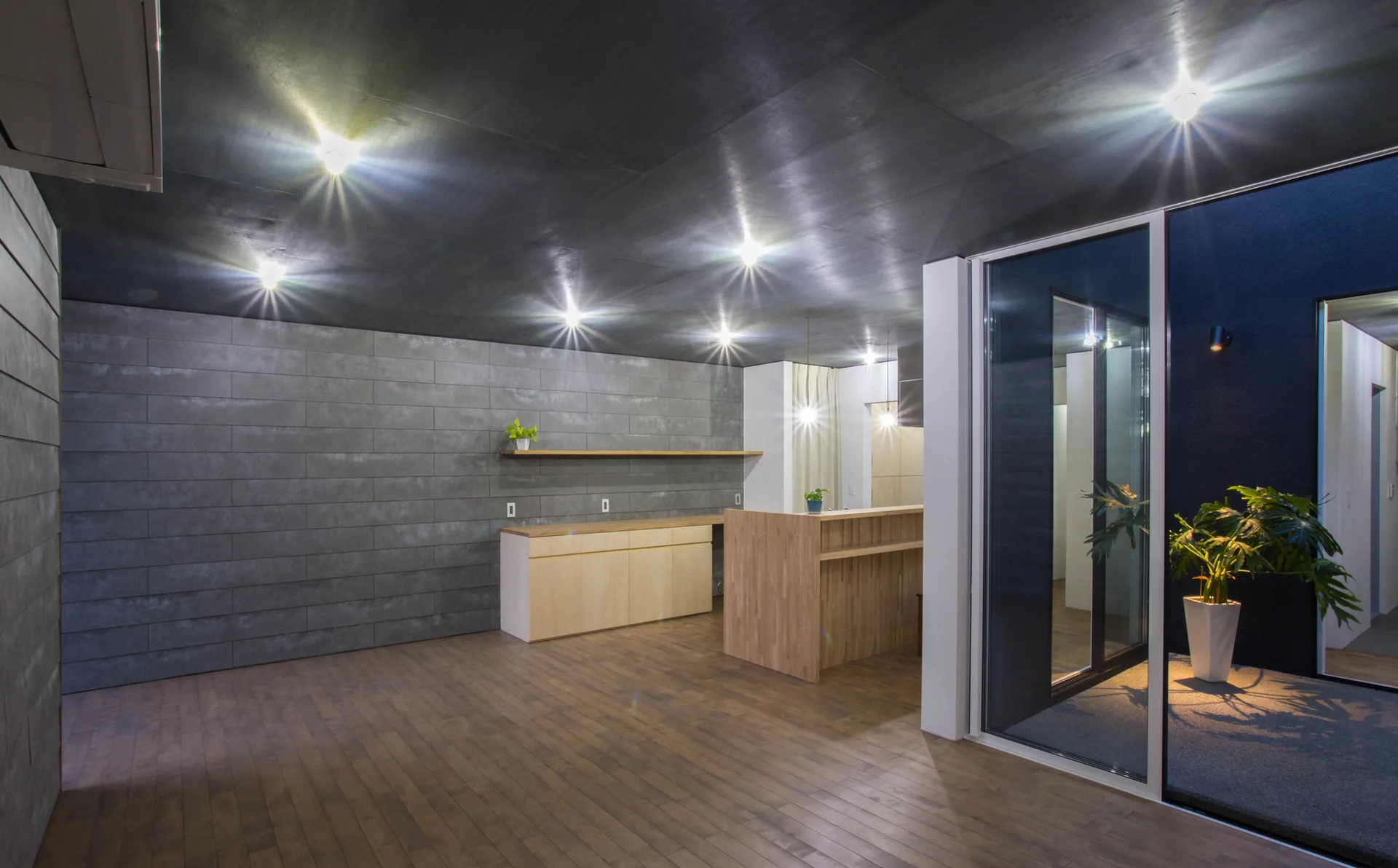 A modern kitchen features a grey tiled feature wall, a light wood island, matching cabinets, a dark ceiling with spotlights, and sliding glass doors.