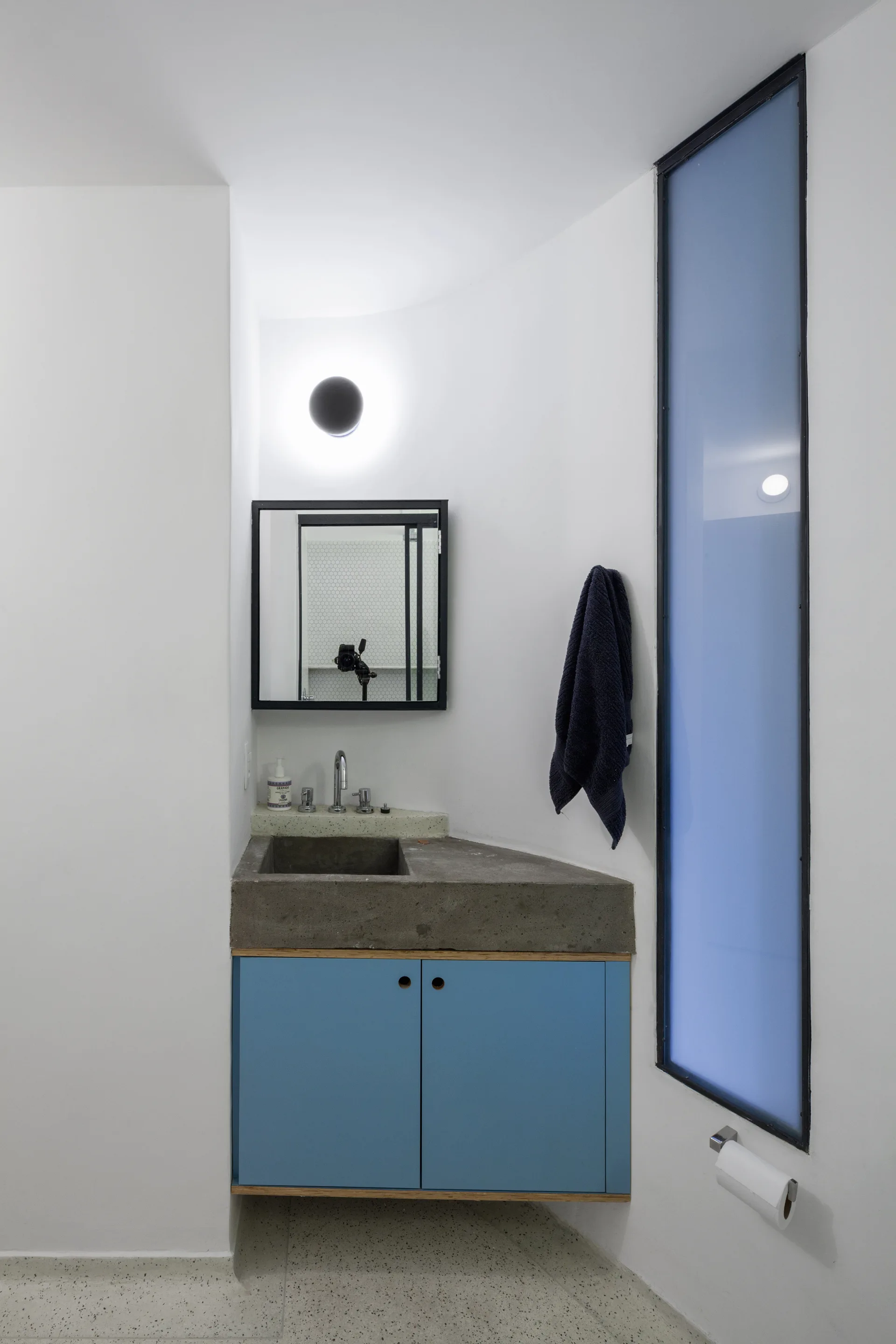 A minimalist powder room features an angled concrete sink, blue vanity cabinets, and a tall tinted window set against white walls.