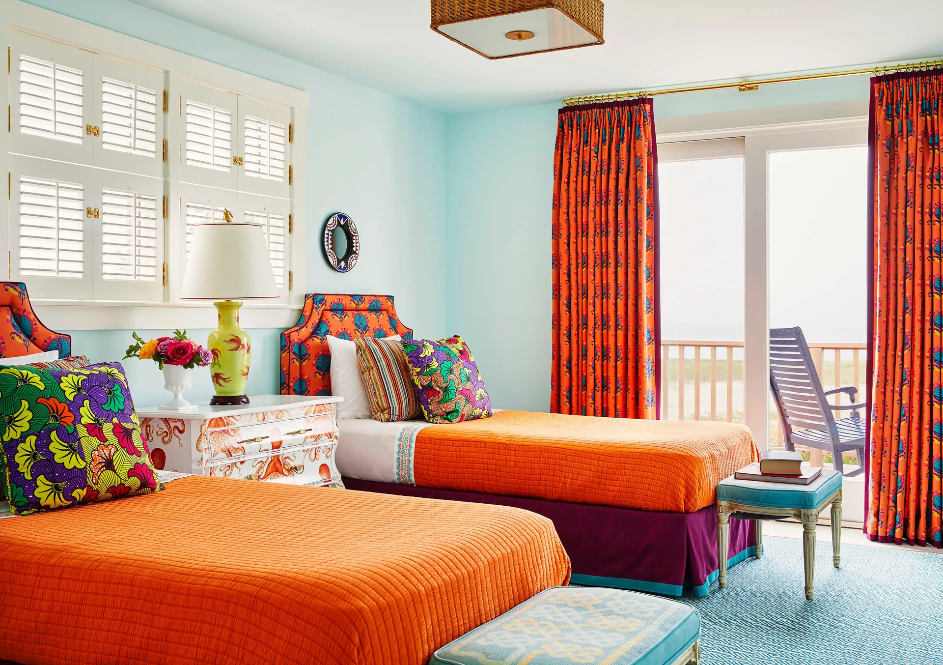 A bright blue bedroom features two twin beds with vibrant orange quilted covers, patterned headboards, and a white nightstand with an octopus illustration.