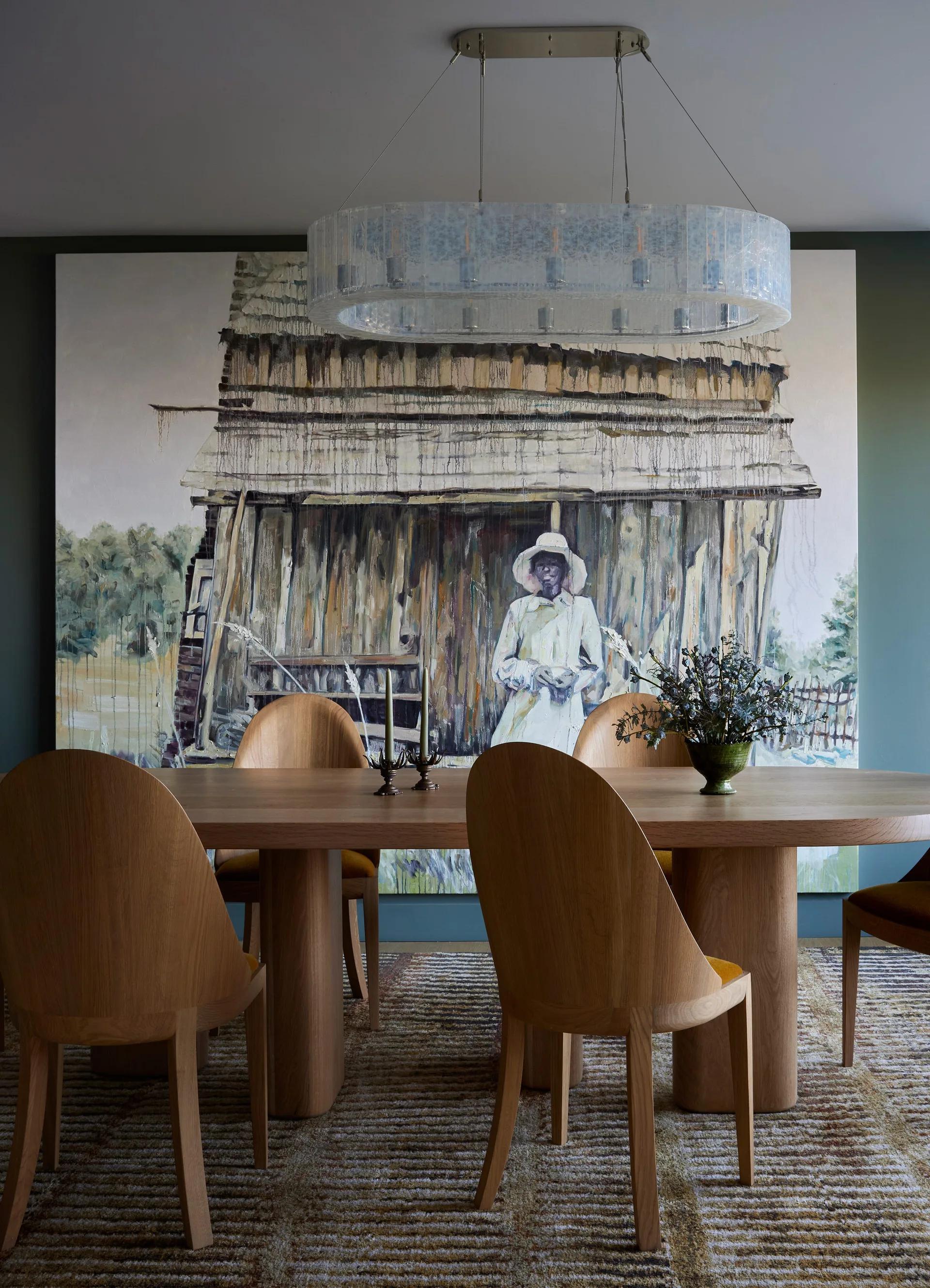 A modern dining room features an oval wooden table, rounded wood chairs, a large textured glass chandelier, and an oversized painting on a deep green wall.