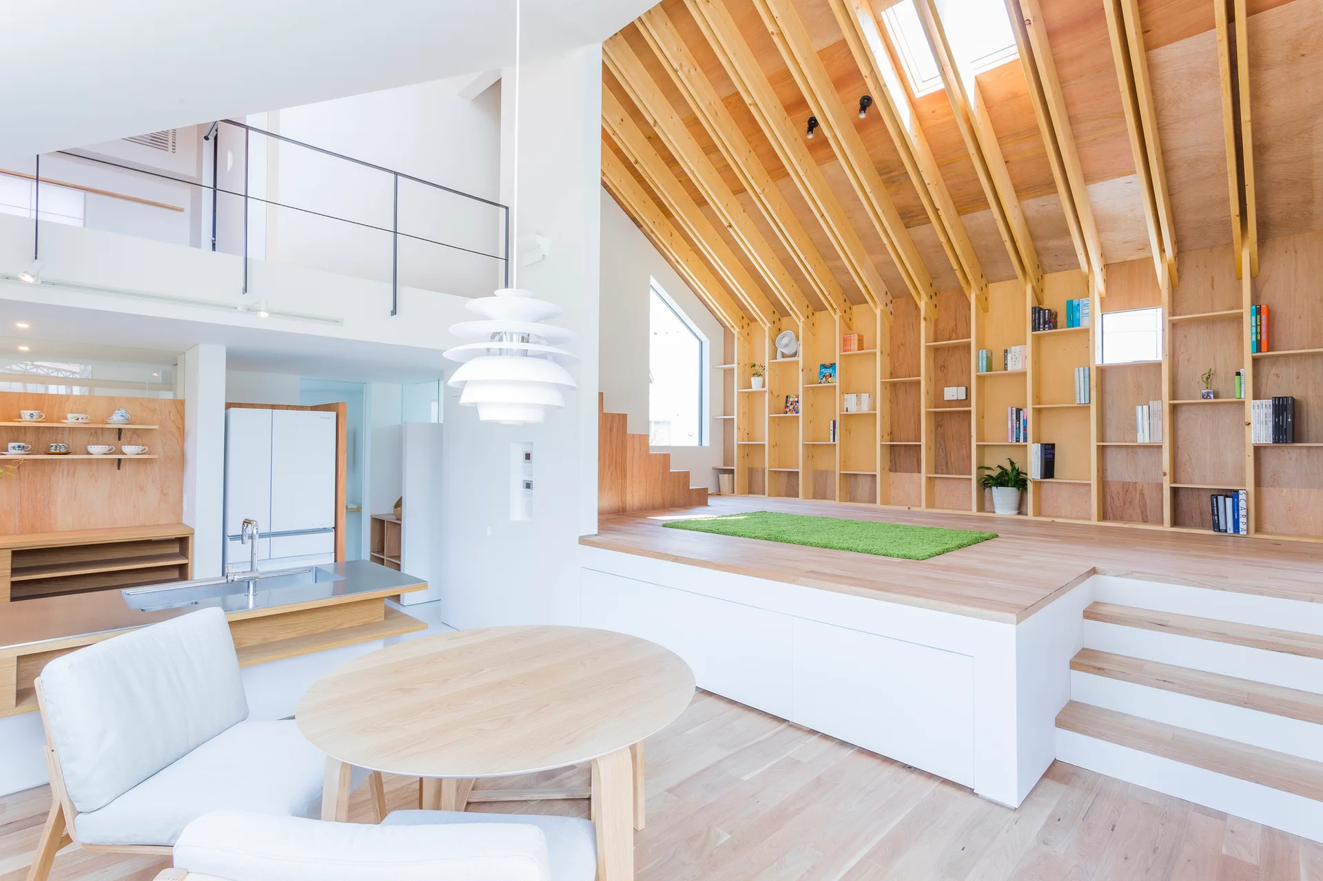 A bright open-plan room features a dining table in the foreground and a raised wooden platform under a vaulted ceiling with integrated shelving.