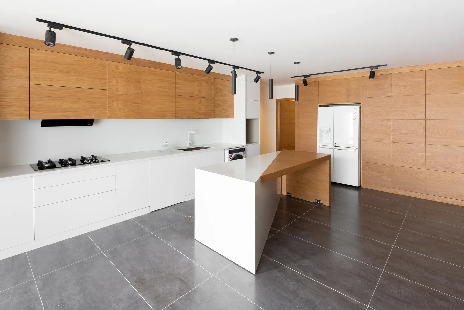 A minimalist kitchen features two-toned white and wood cabinets, an asymmetric island with an integrated wood bar, and dark grey tile flooring.