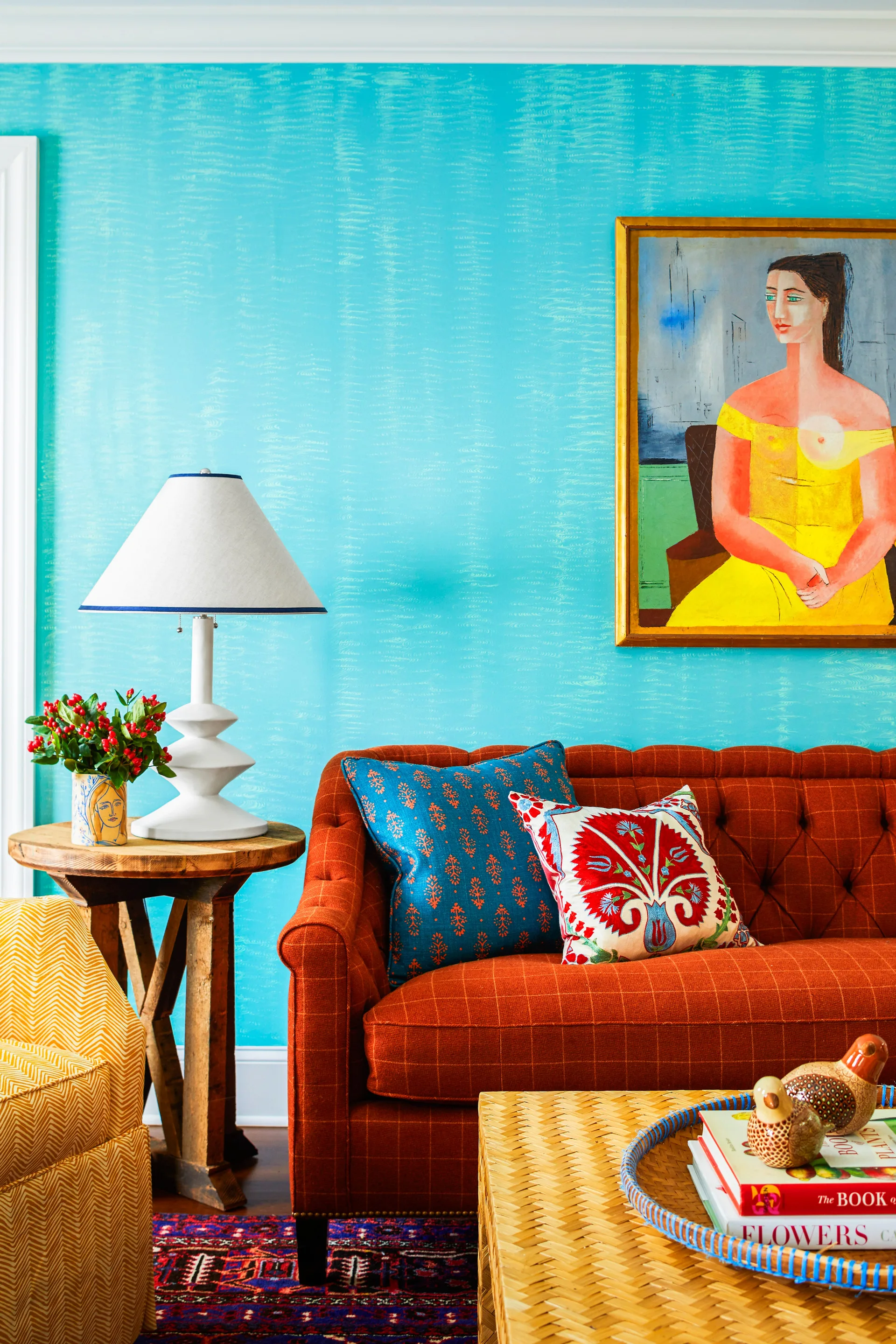 A tufted orange sofa with patterned pillows sits against a textured blue wall next to a white table lamp on a wooden side table.