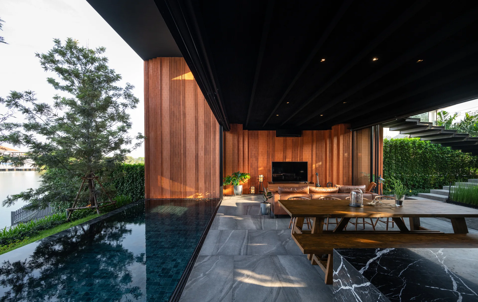 A modern indoor-outdoor living space with wood-slatted walls, grey stone floor tiles, a tan leather sofa, and an integrated swimming pool.