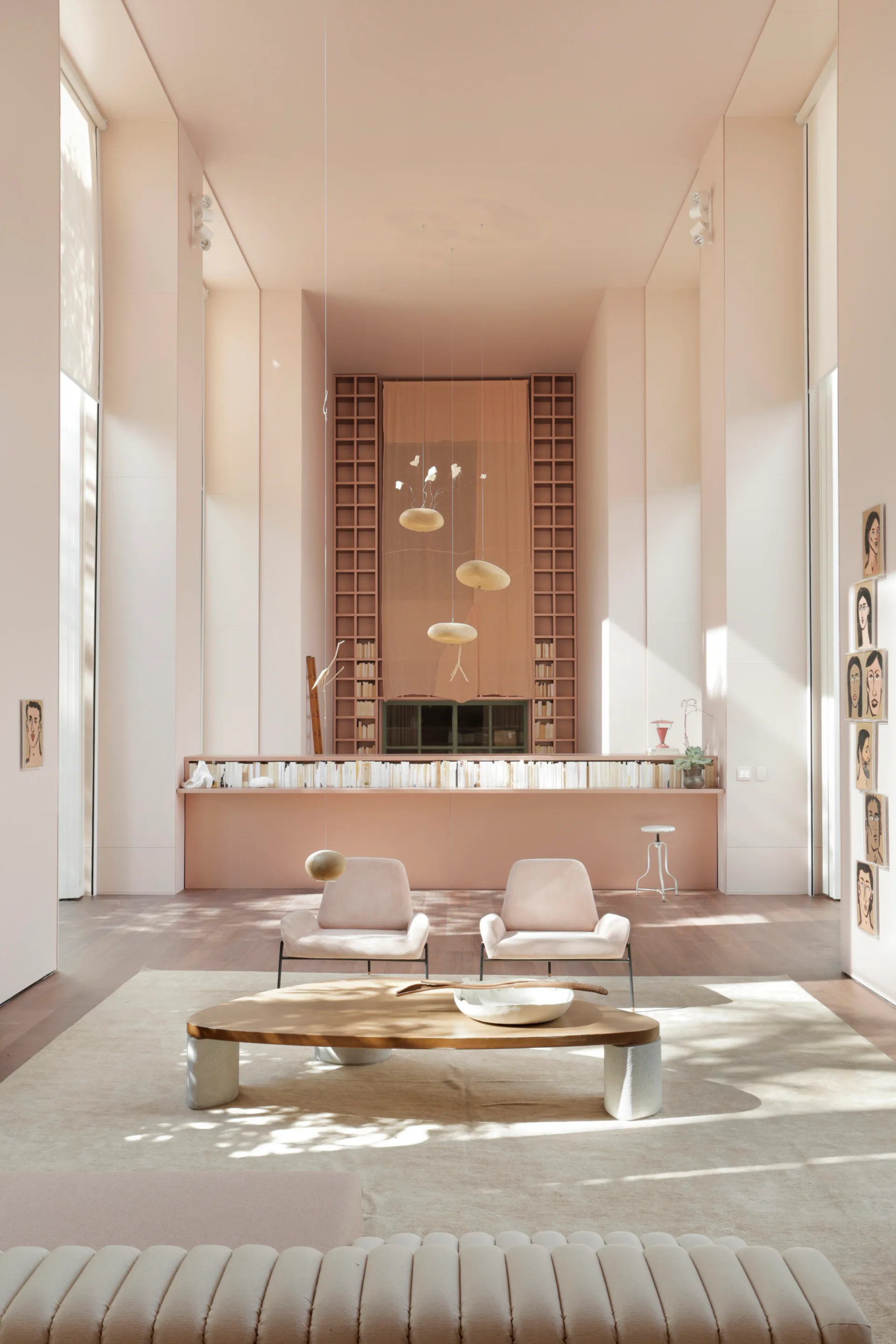 A high-ceilinged blush pink living room features two minimalist armchairs, an organic-shaped wooden coffee table, and tall built-in bookshelves.