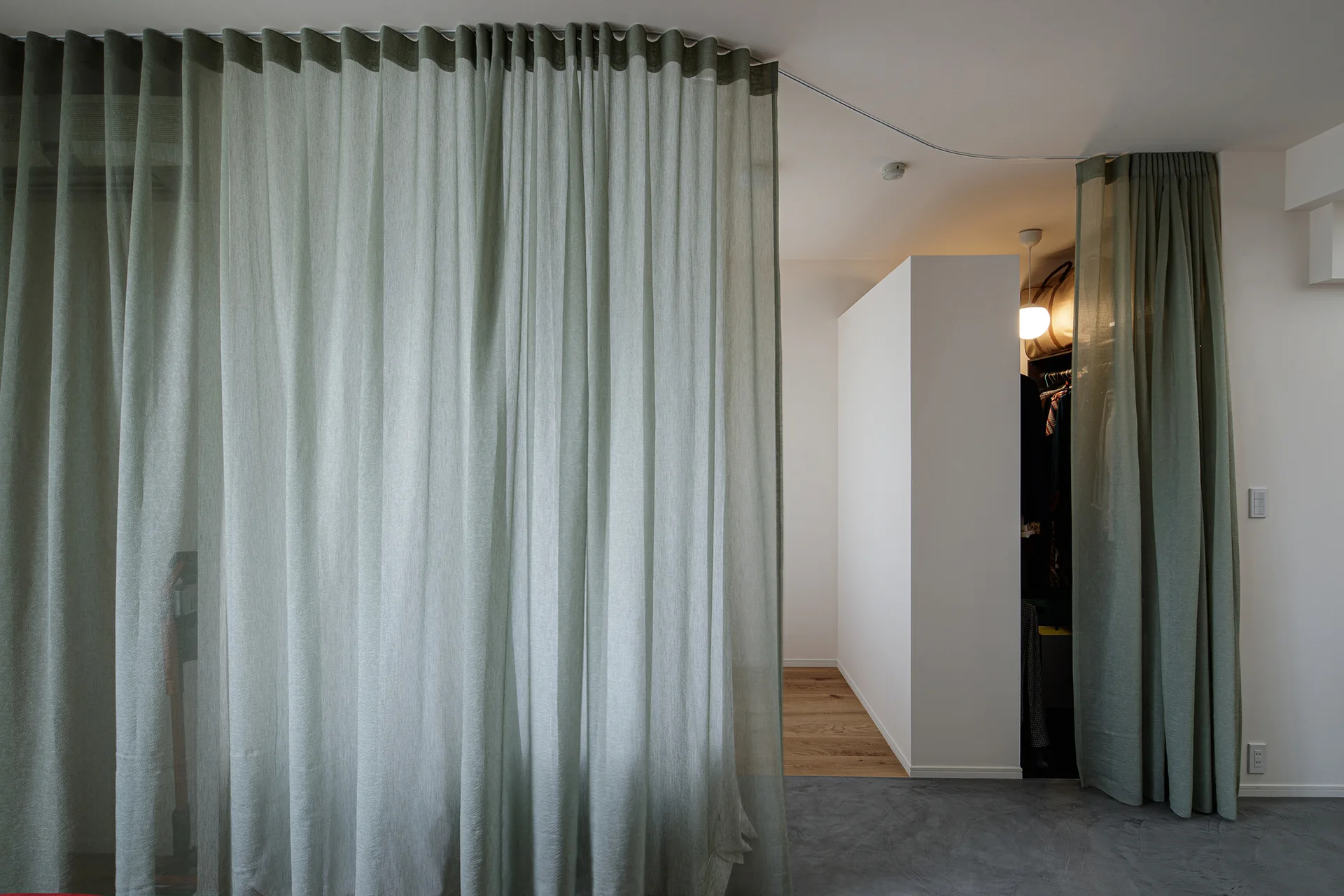 A floor-to-ceiling sage green sheer curtain divides a minimalist room from a dressing area with a white partition wall and grey concrete floor.
