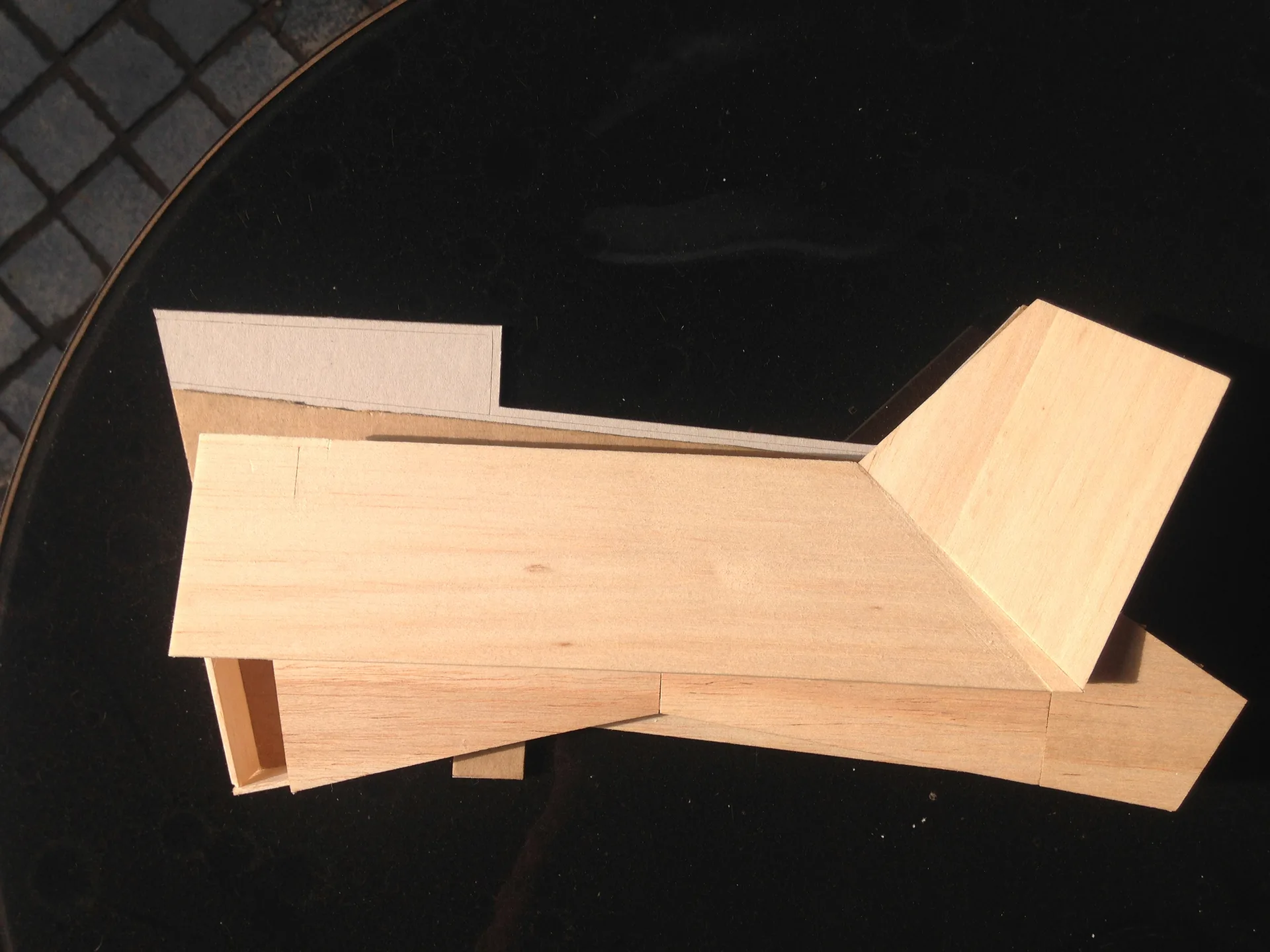 A balsa wood architectural study model with angular planes rests on a black reflective surface in bright sunlight.