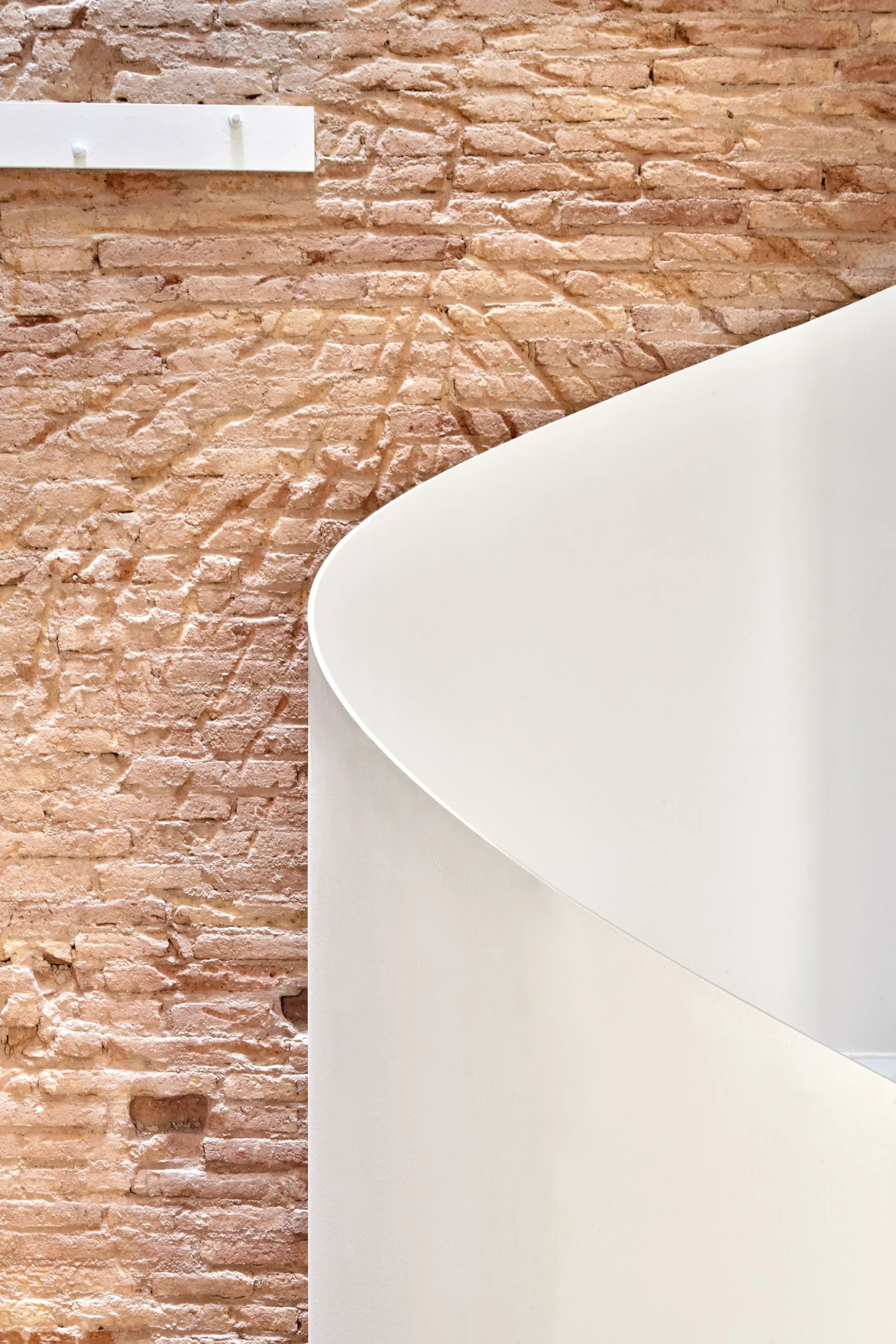 A smooth white curved staircase railing contrasts against a textured rustic orange brick wall with a small white metal cleat mounted on top.