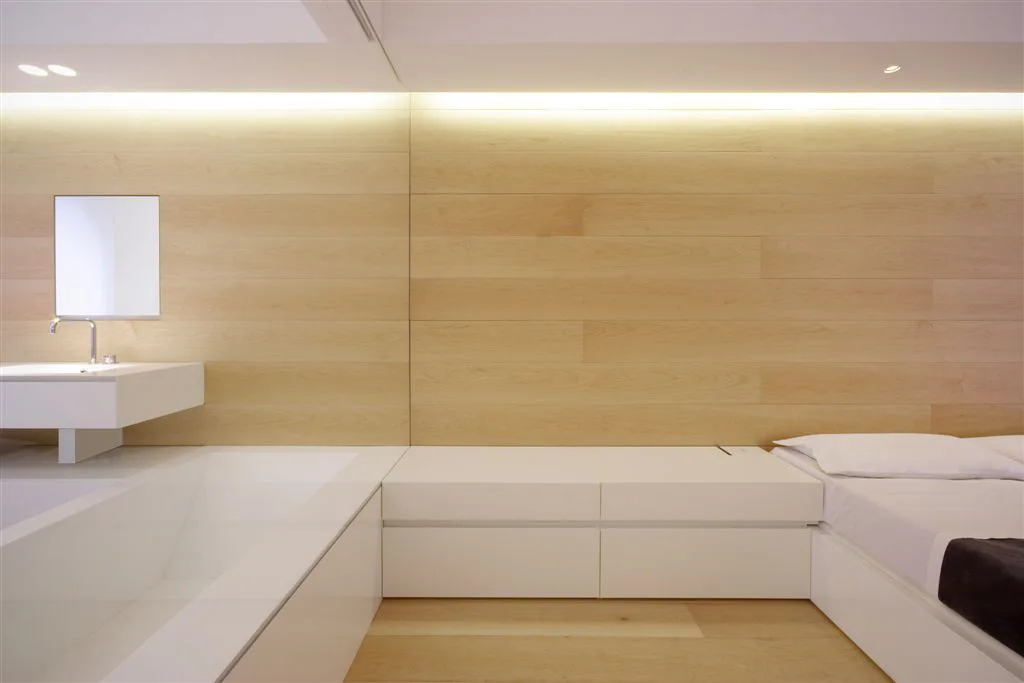 A minimalist bedroom and open bathroom suite featuring light wood wall paneling, white built-in platform storage, a bathtub, and cove lighting.