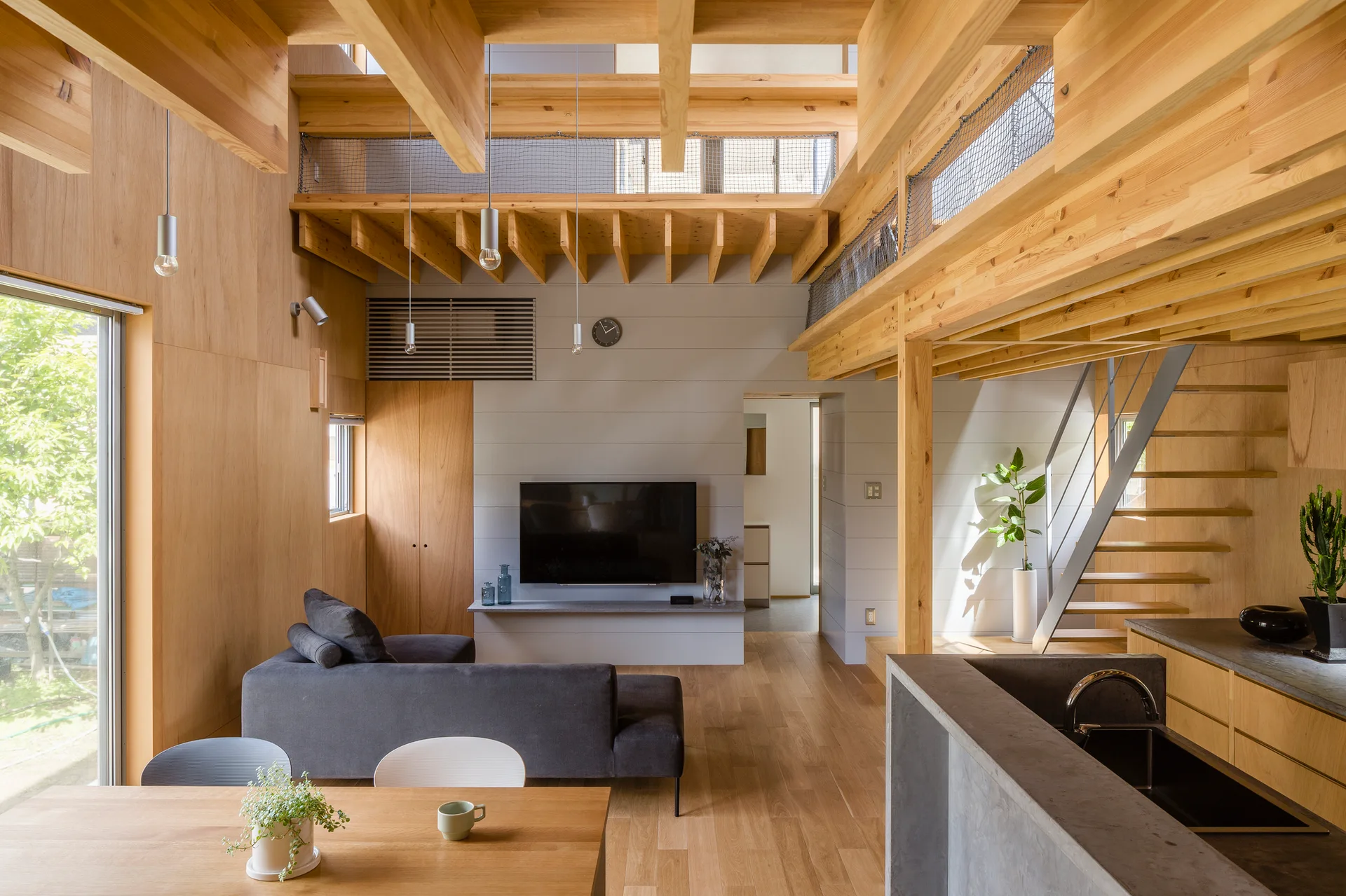 A Japanese modern living room featuring a grey sofa, wall-mounted television, wooden staircase, and exposed structural timber beams.