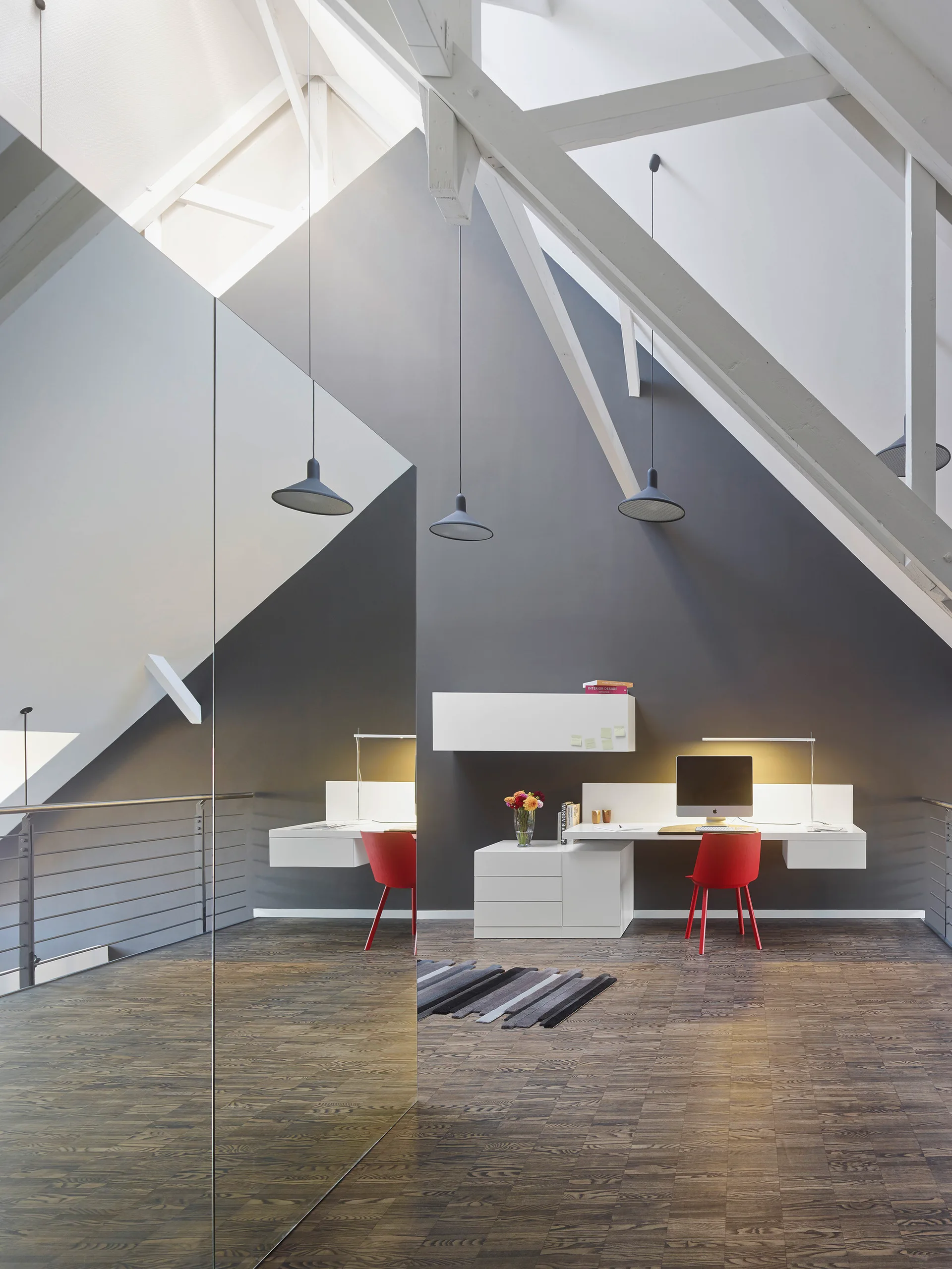A loft home office featuring a white wall-mounted desk, two bright red chairs, a dark grey accent wall, and exposed white ceiling beams.