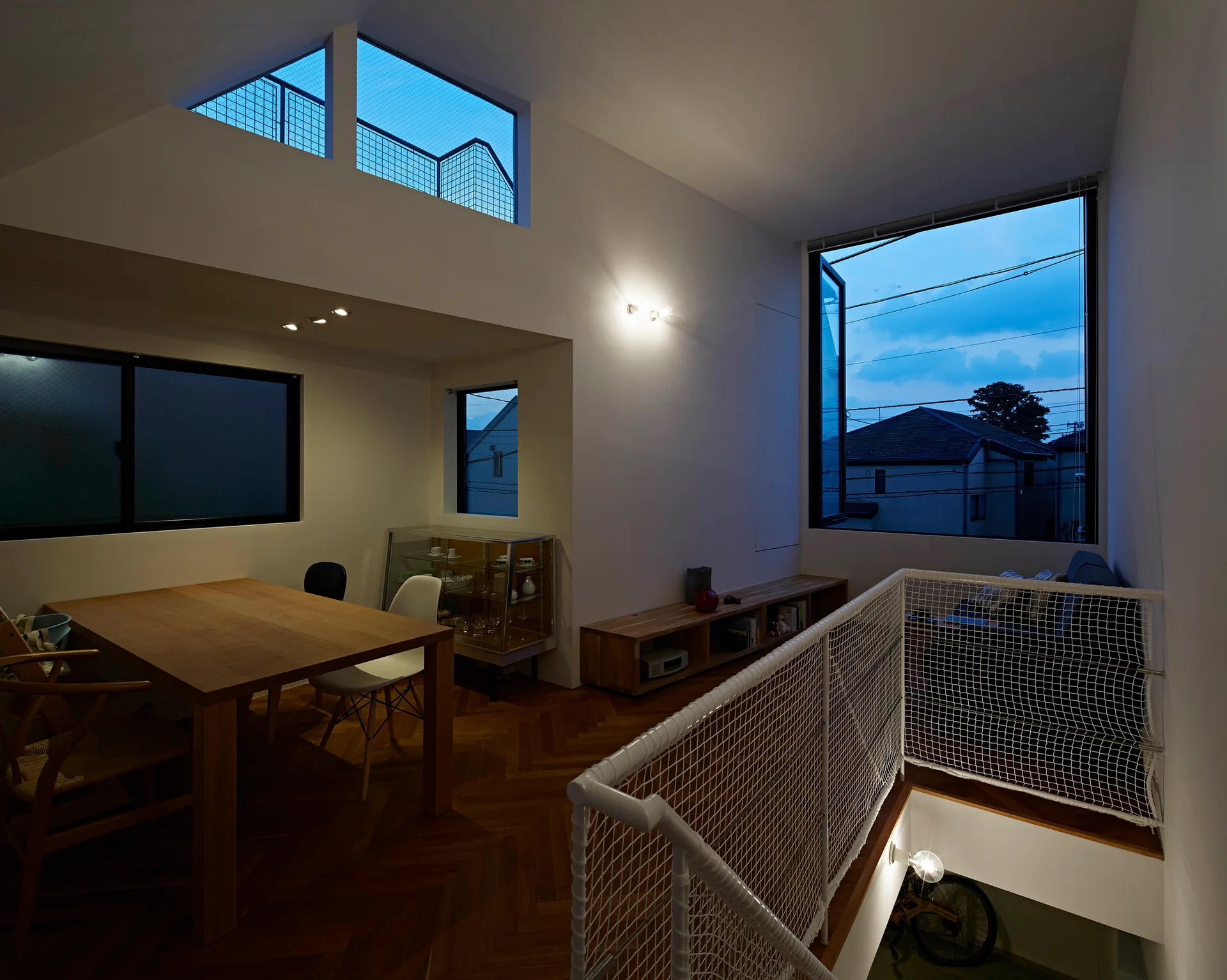 A modern mezzanine dining room features a wooden table, herringbone parquet flooring, and a white safety net railing overlooking the stairs.