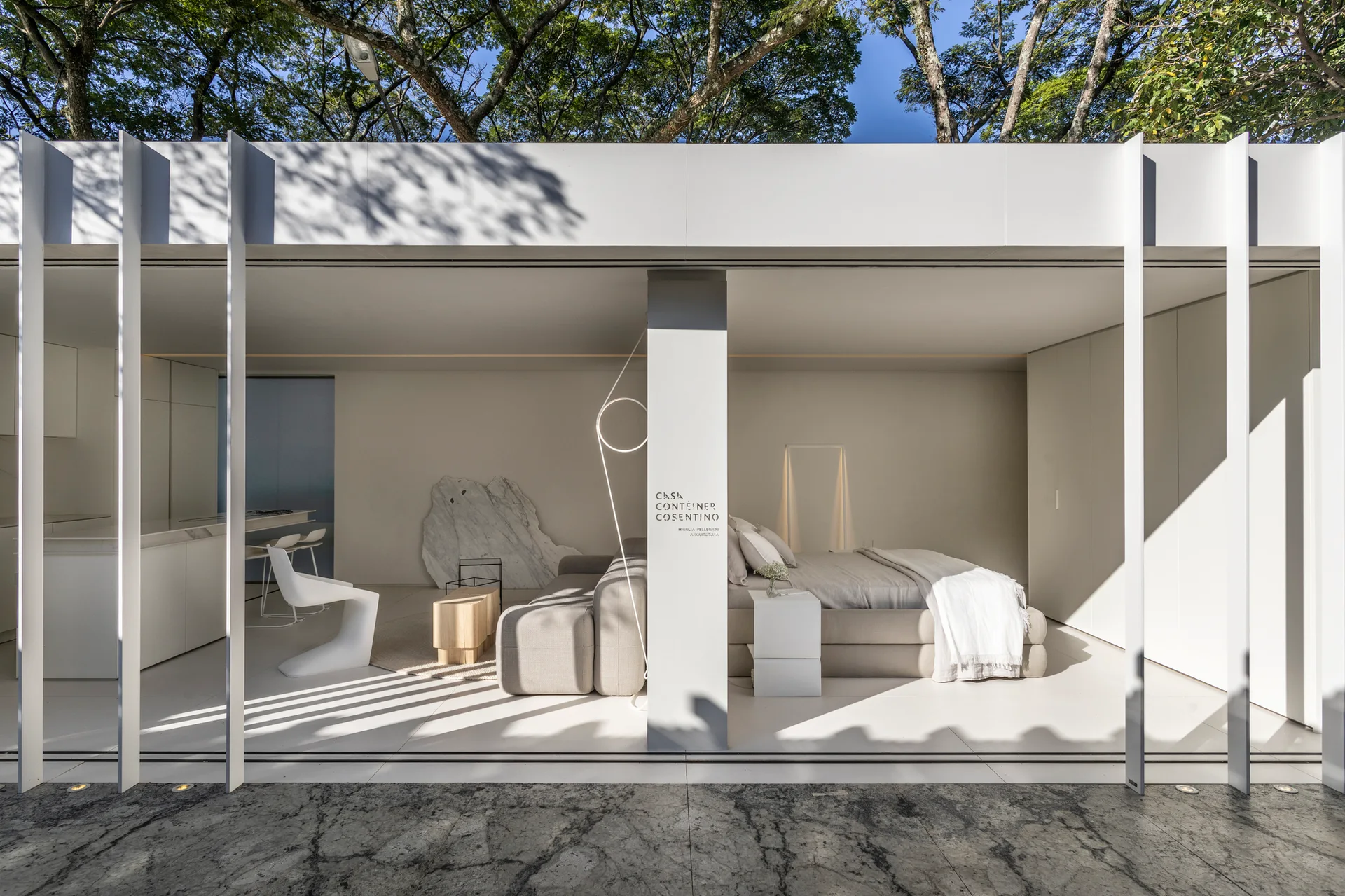 An open-concept bedroom and living space features a gray modular sofa, white platform bed, and clean white tile floors open to the outdoors.