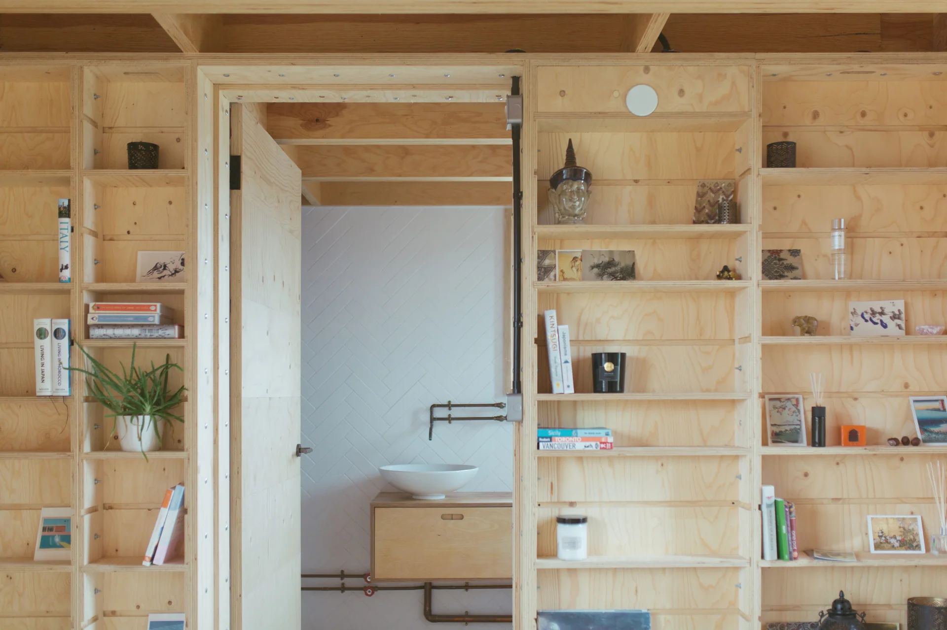 A light plywood shelving unit frames a doorway into a small bathroom featuring a white vessel sink and a tiled herringbone wall.