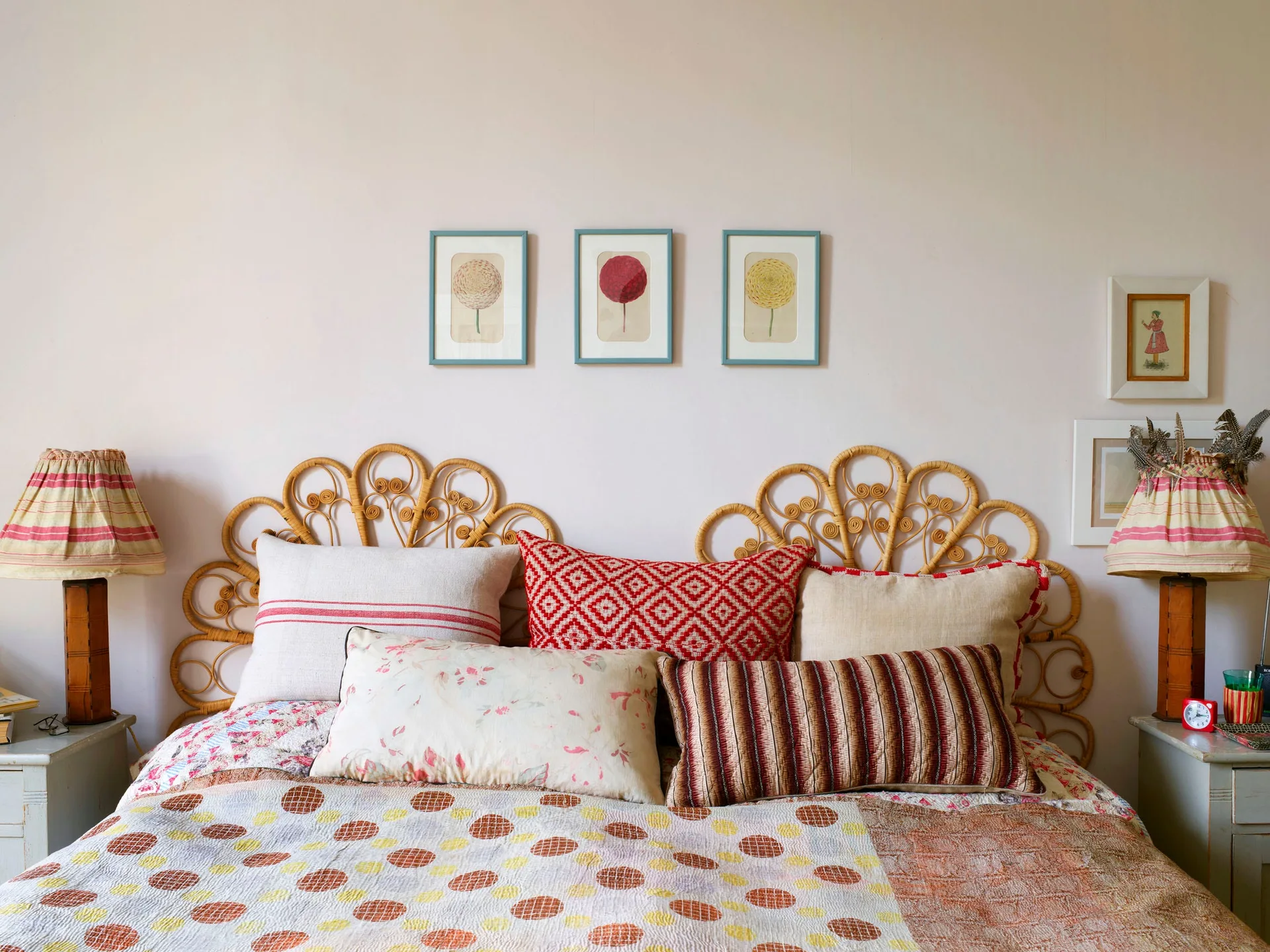 An eclectic bedroom features a scalloped rattan headboard, a bed layered with patterned pillows, and small framed artwork hanging on an off-white wall.