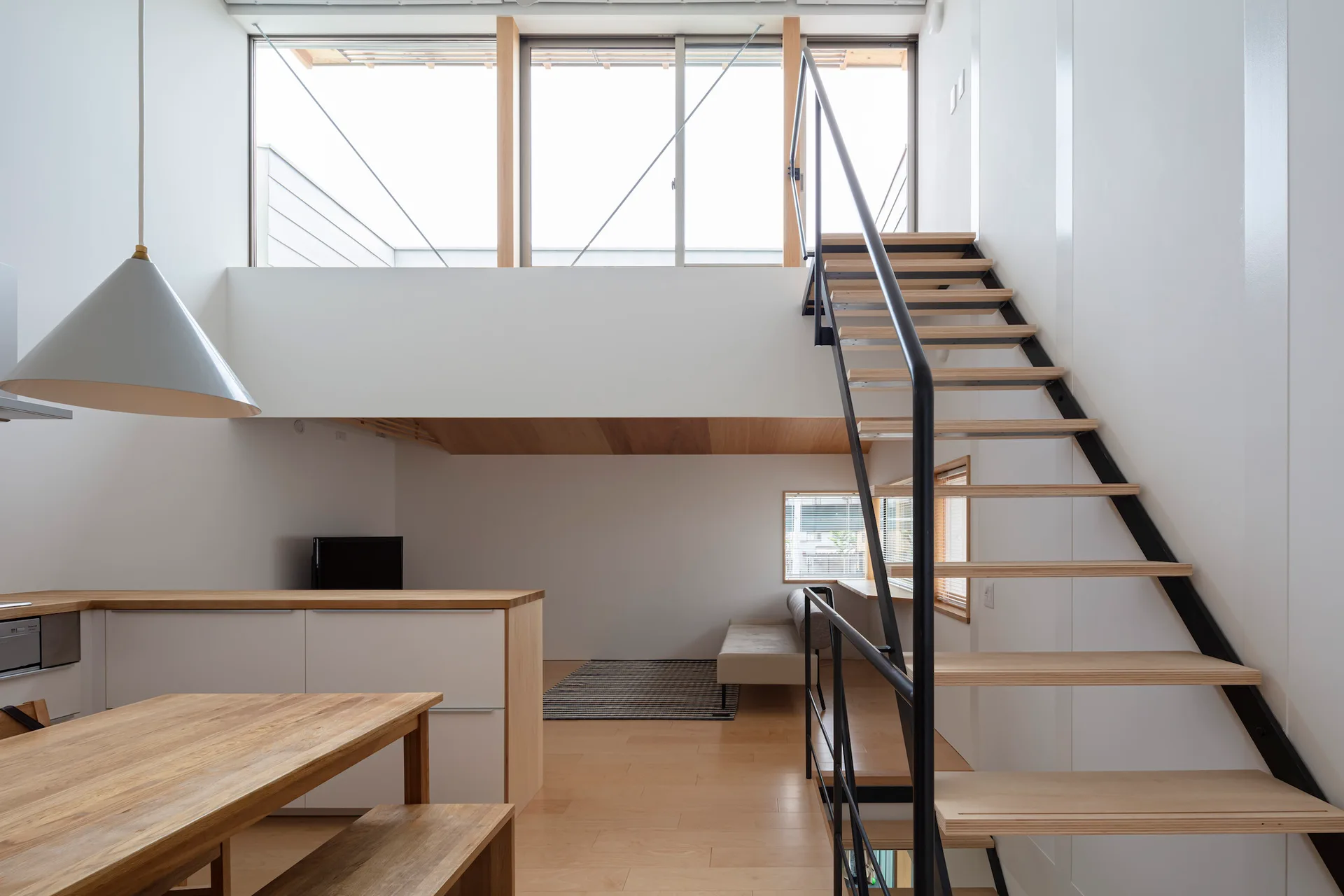 A bright minimalist home features an open riser wood and steel staircase leading to a mezzanine, alongside a wood-topped kitchen island and dining table.