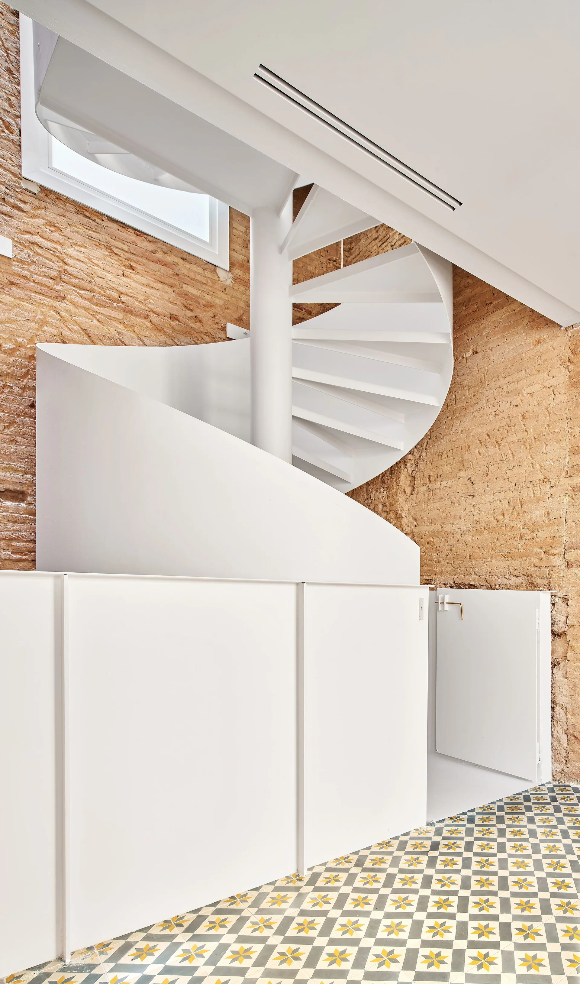 A white metal spiral staircase curves upward against an exposed terracotta brick wall above geometric patterned floor tiles and built-in white cabinets.