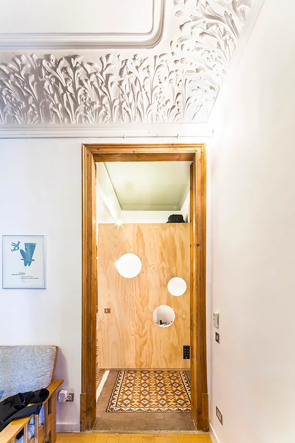 A view through a wooden doorway showing an ornate plaster ceiling, patterned floor tiles, and a plywood wall panel with circular lights.