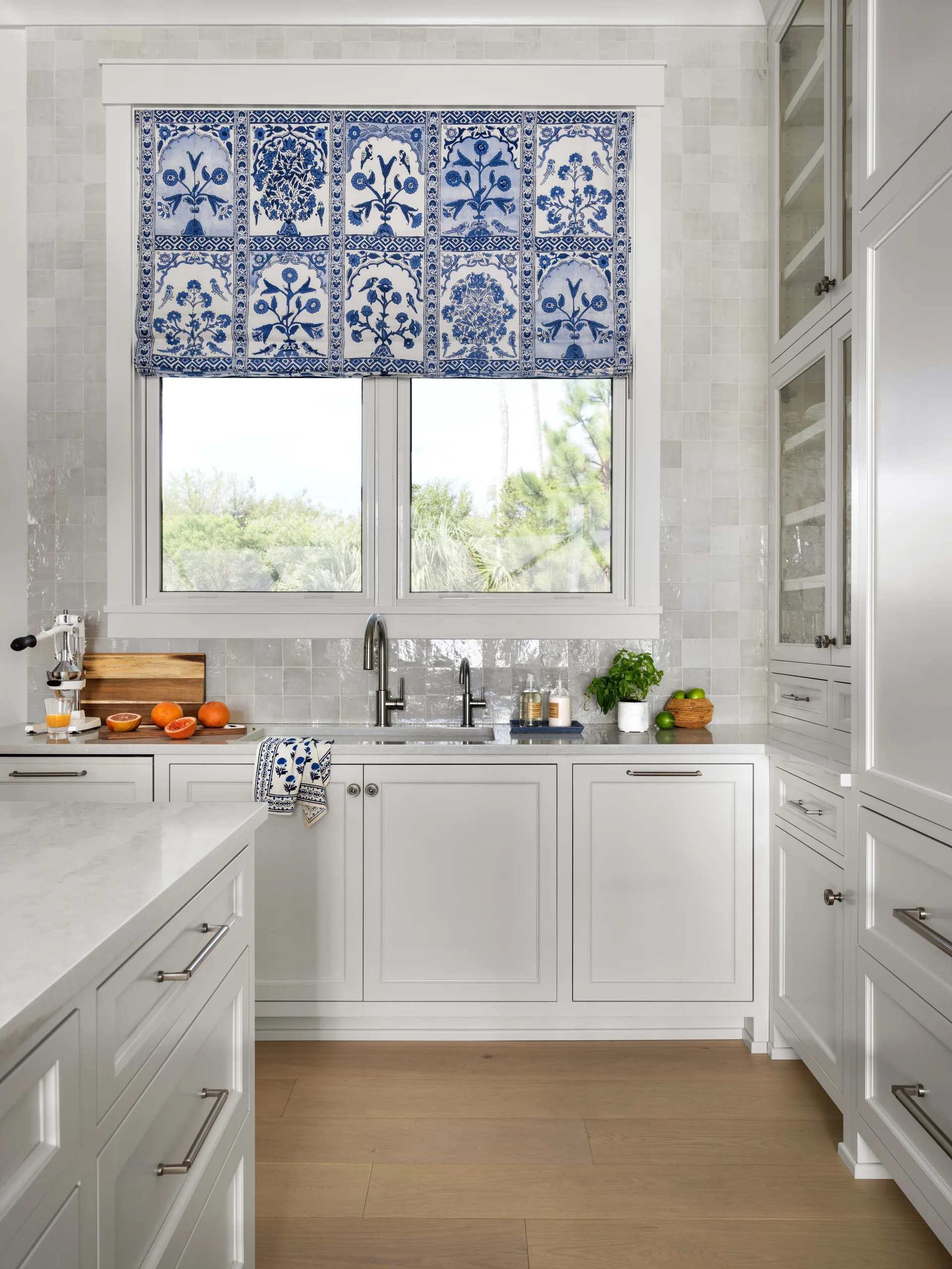 A kitchen sink area features white cabinetry, a glossy square tile backsplash, and a blue and white botanical patterned roman shade on the window.