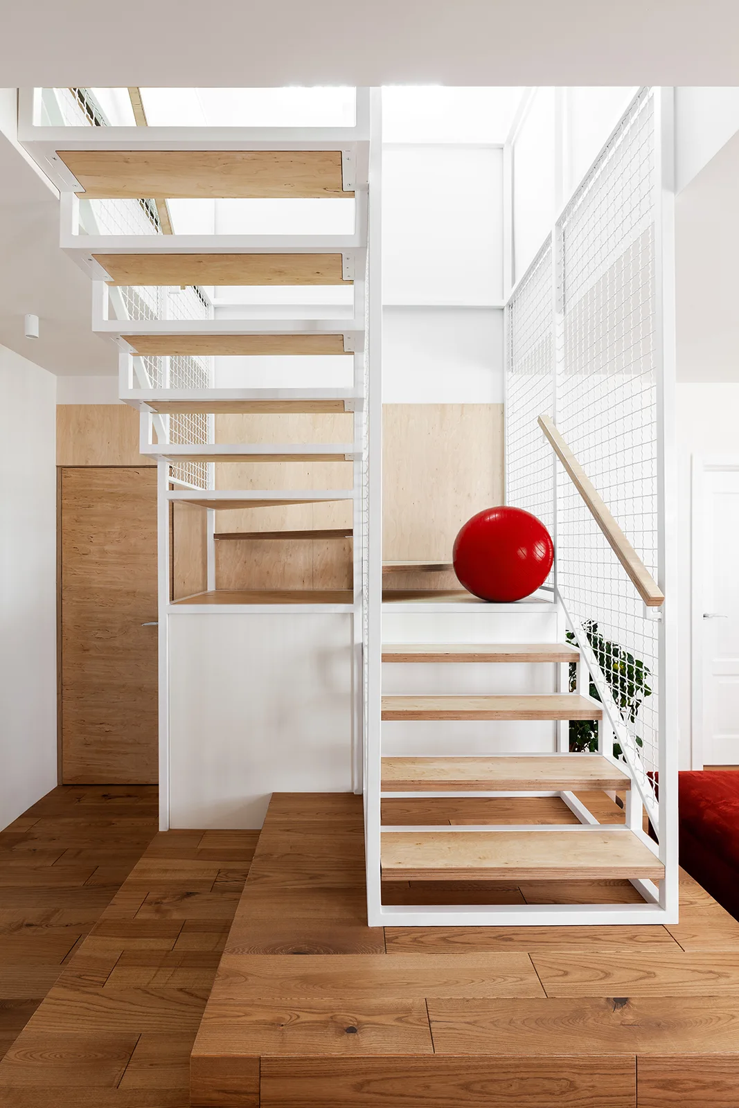 A modern white metal staircase features open light wood treads and a white mesh wire railing set against white walls and a light wood door.