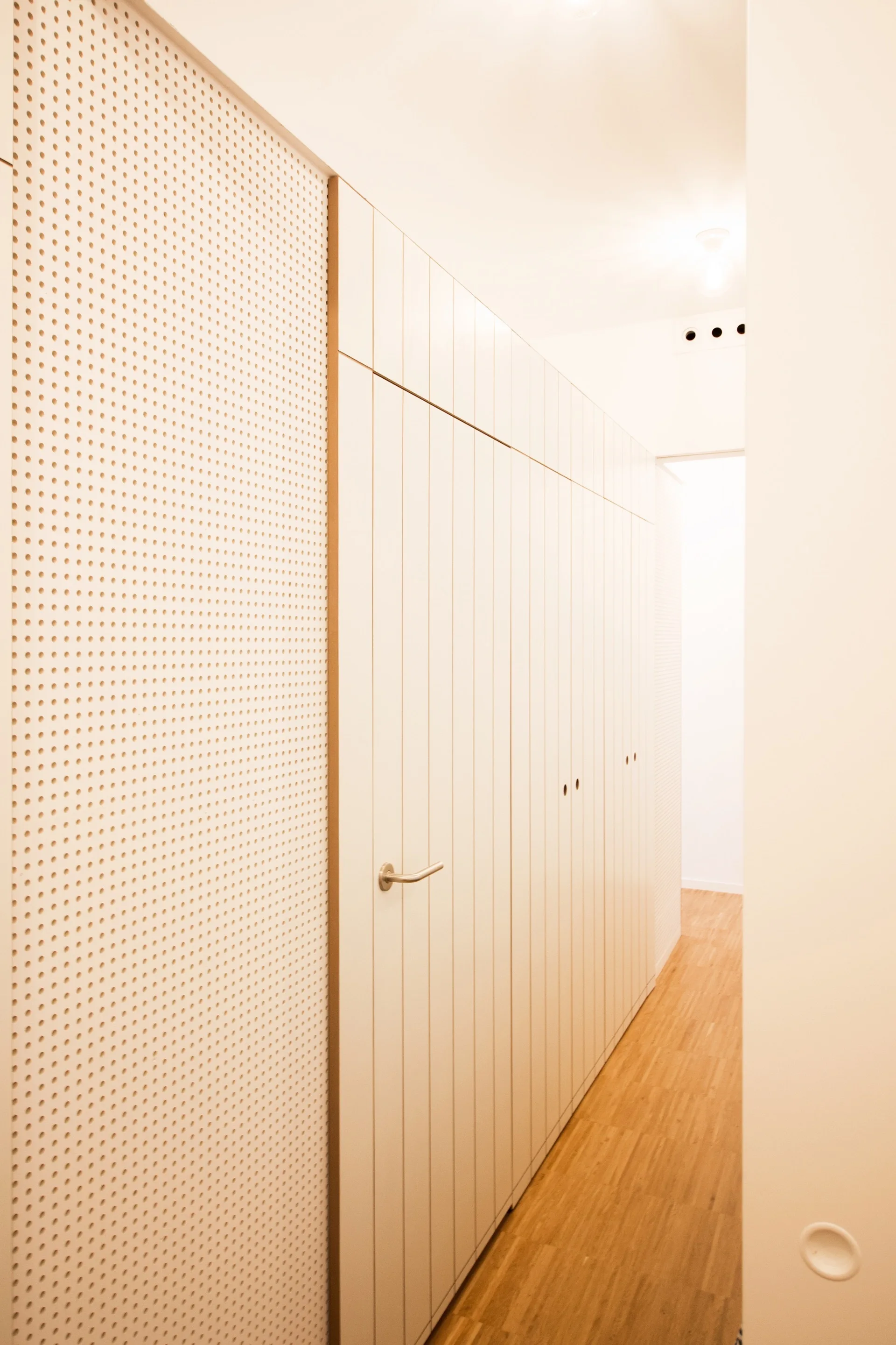 A minimalist hallway featuring tall white built-in closets with vertical paneling next to a perforated white accent wall and light wood flooring.