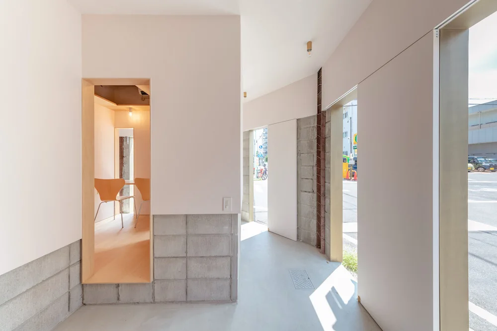 A minimalist entryway features smooth white walls above concrete cinder blocks, a light wood framed rectangular opening, and a concrete floor.