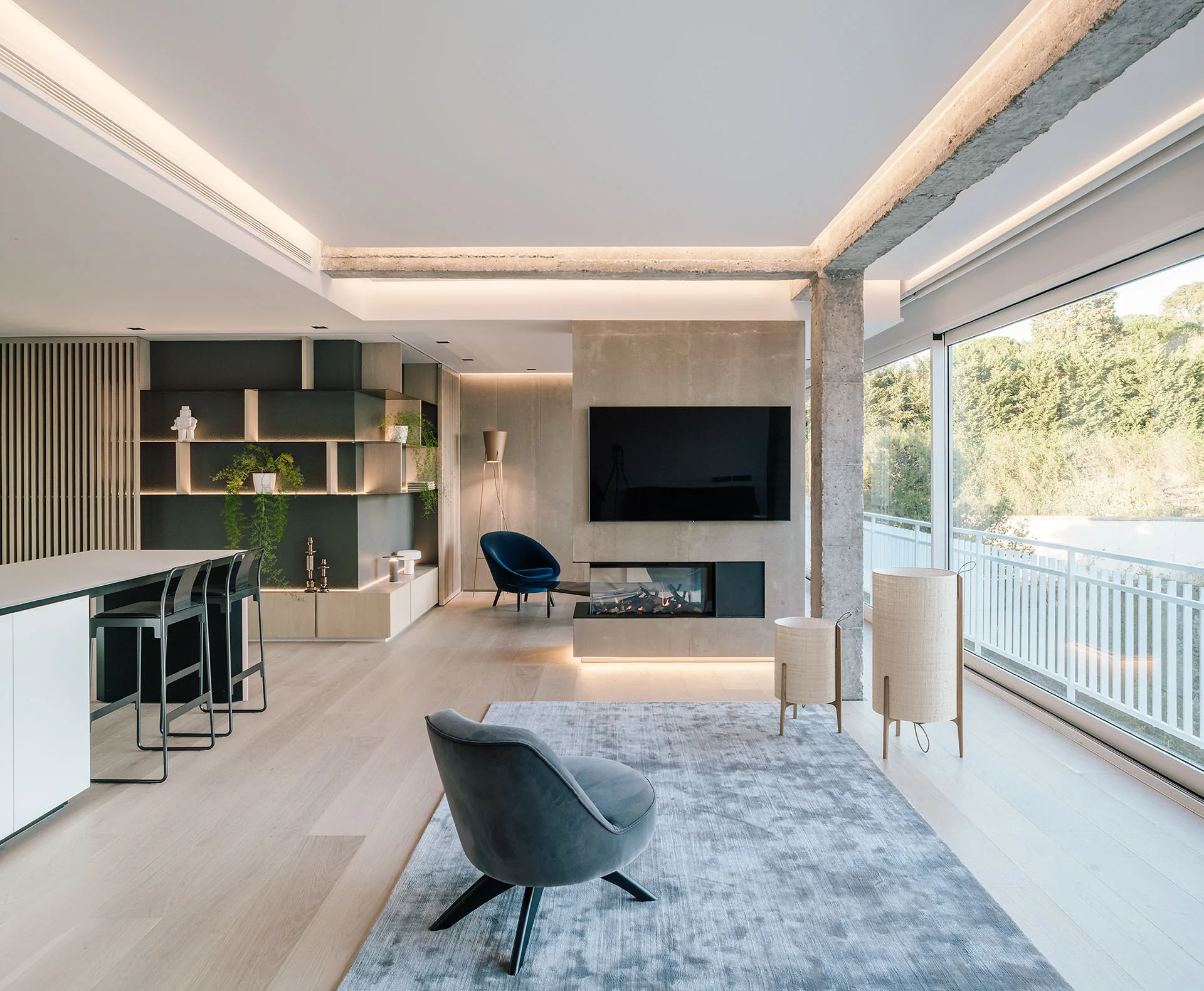 A modern open-plan living room features a built-in concrete fireplace, a gray upholstered armchair, exposed concrete columns, and large windows.