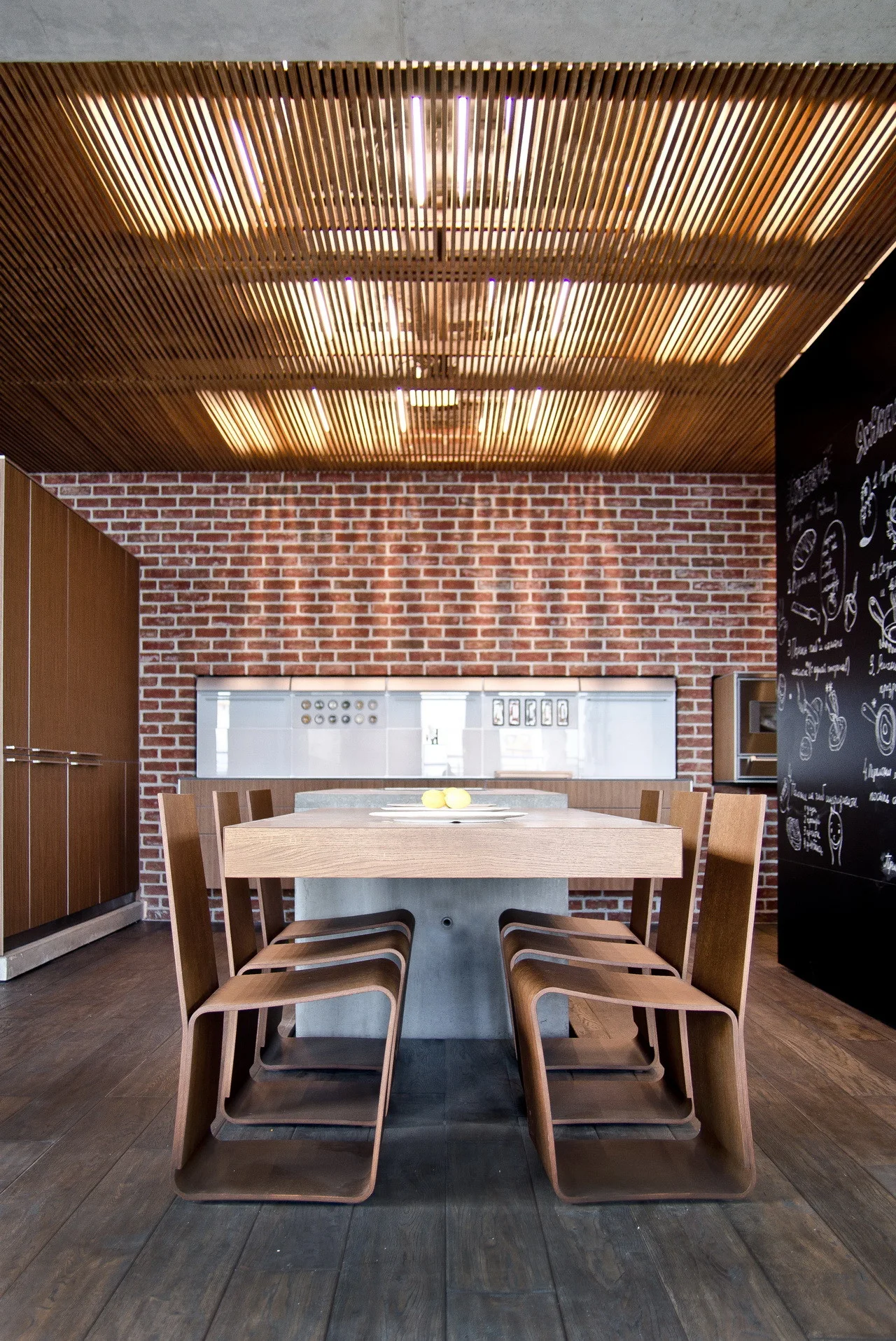 A modern kitchen dining space features a wood dining table and curved chairs against an exposed brick wall under a lit wood slat ceiling.
