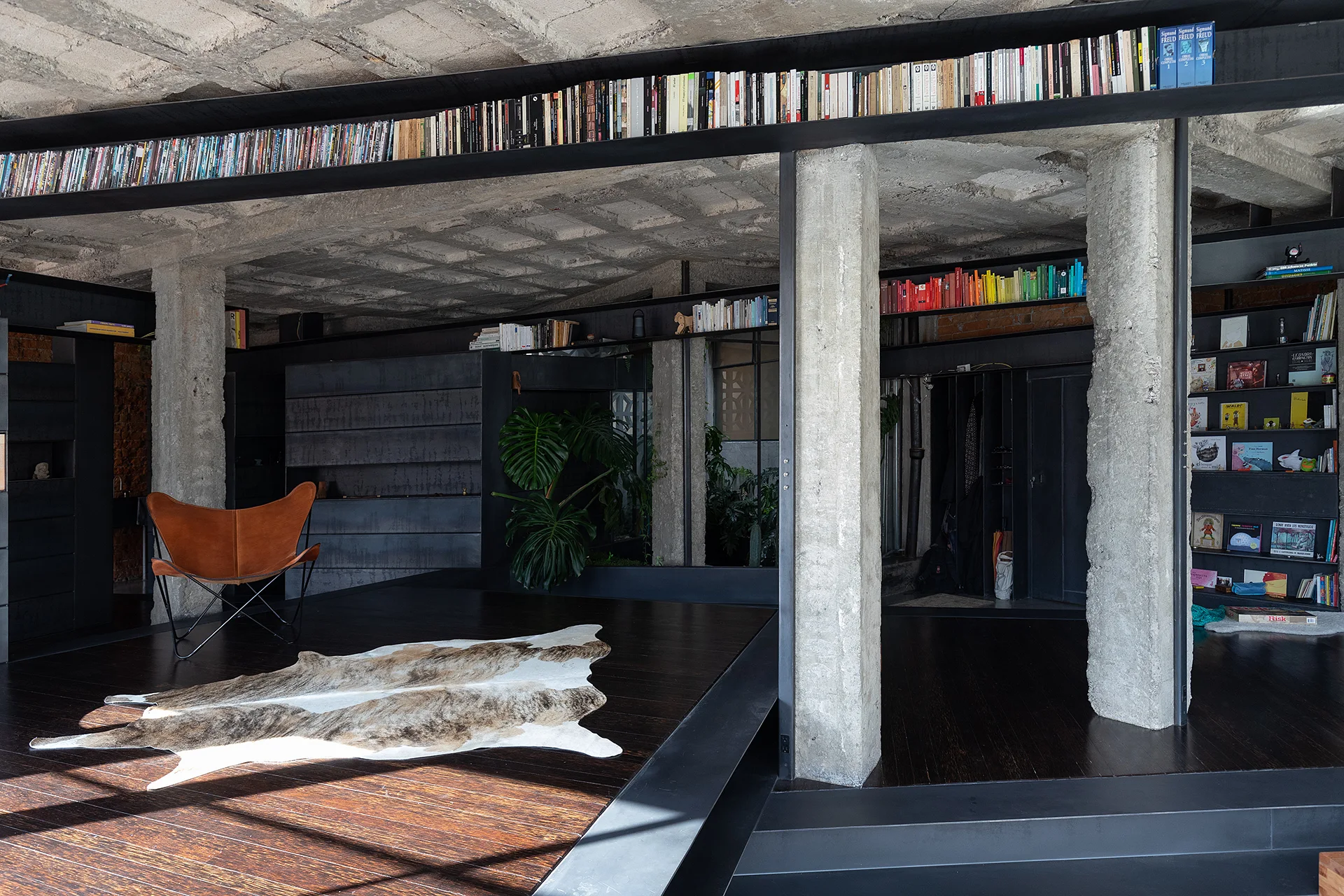 A brutalist living area features raw concrete columns, dark wood floors, a leather butterfly chair on a cowhide rug, and high-level perimeter bookshelves.