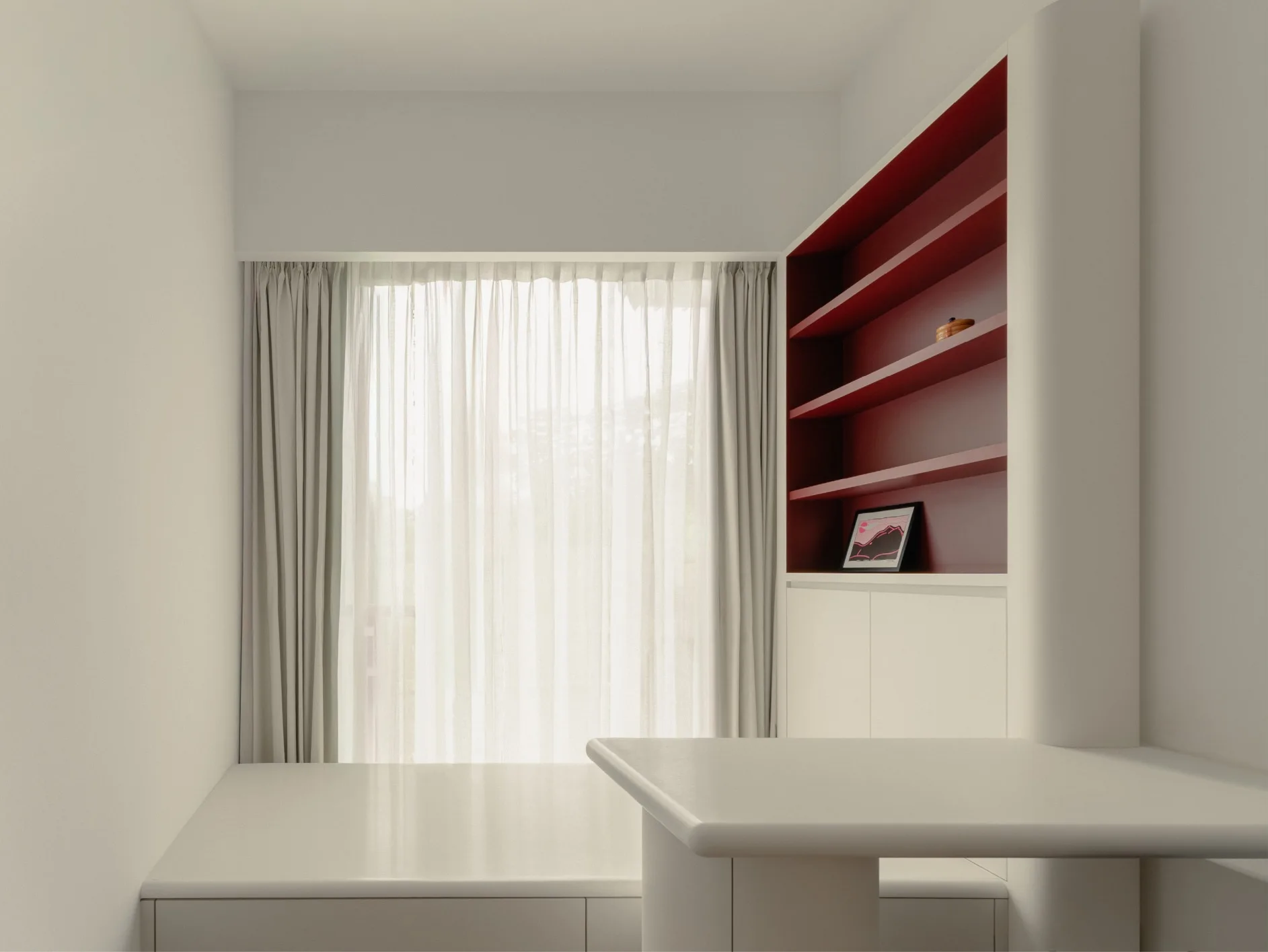A minimalist study nook features an off-white desk, a raised platform, sheer white window curtains, and built-in shelving with a deep red interior.