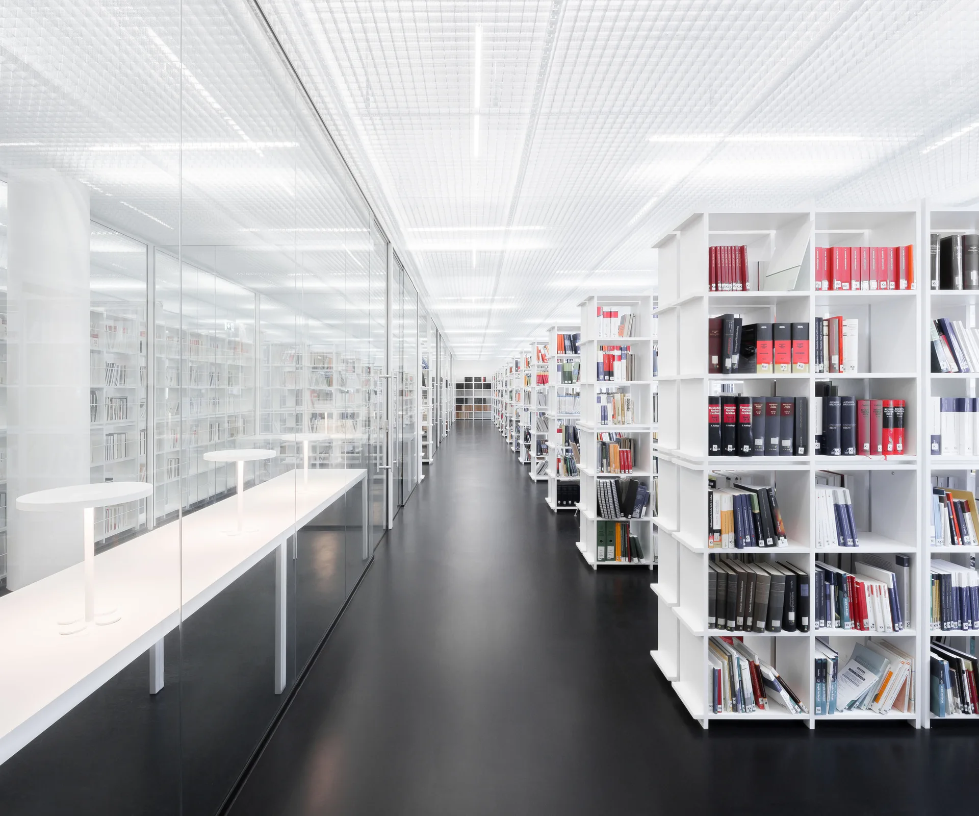 A bright modern library interior features rows of white bookshelves and study desks enclosed in a glass partition under a white grid ceiling.