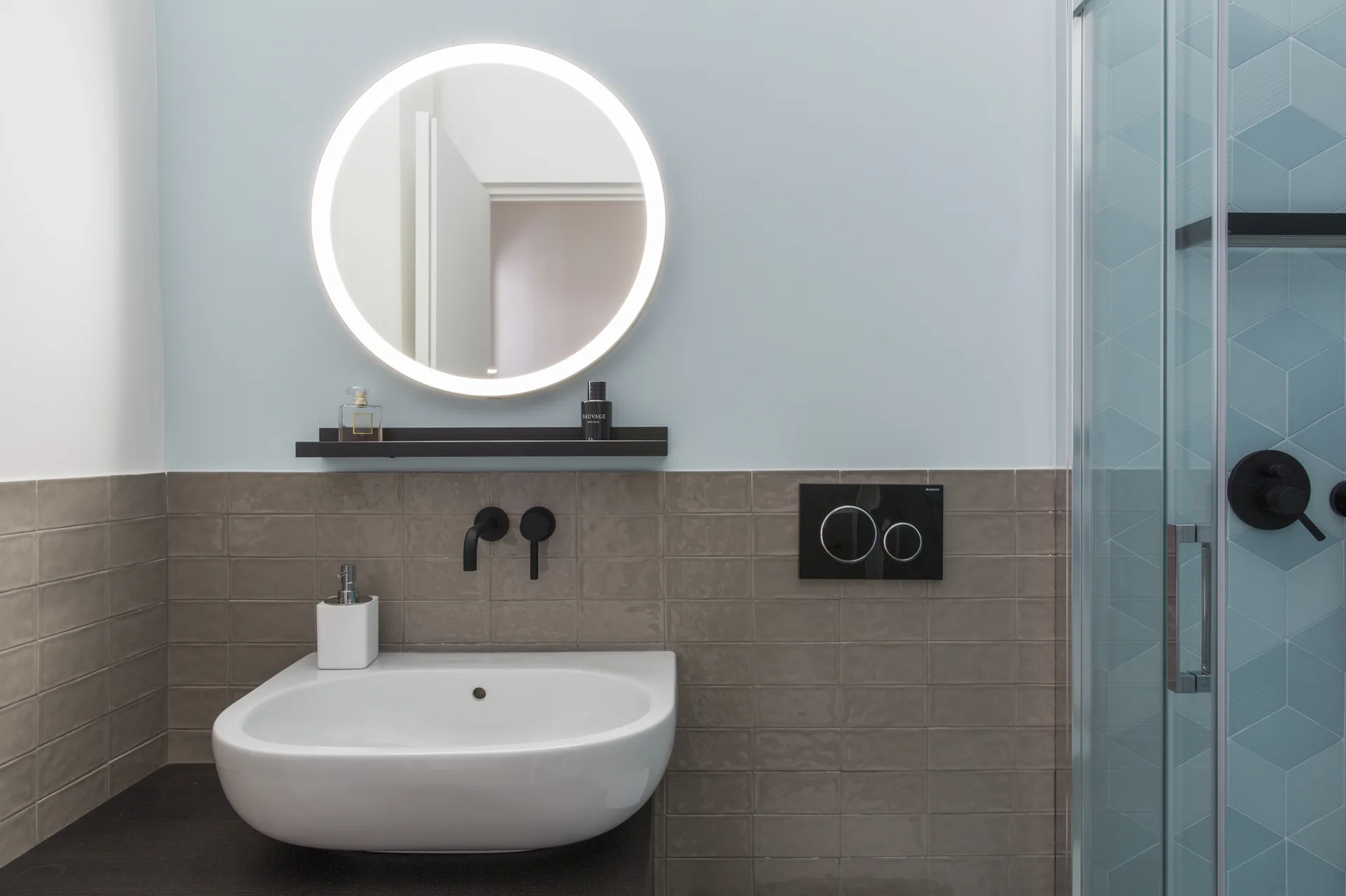 A modern bathroom features a round backlit mirror above a white basin sink, black wall-mounted fixtures, taupe tiles, and a glass shower door.