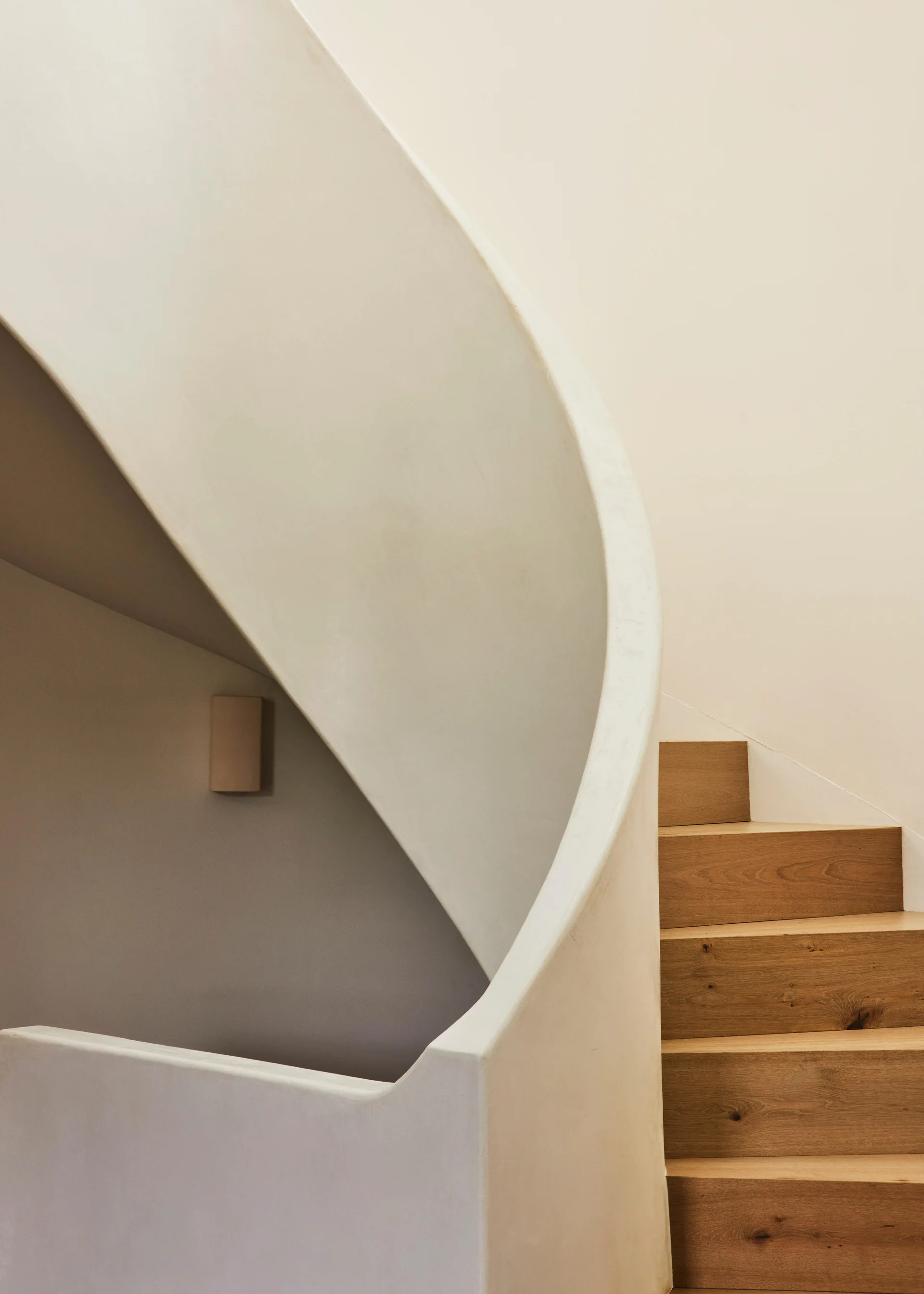 A minimalist curved staircase with smooth off-white plastered walls wrapping around natural wood treads.