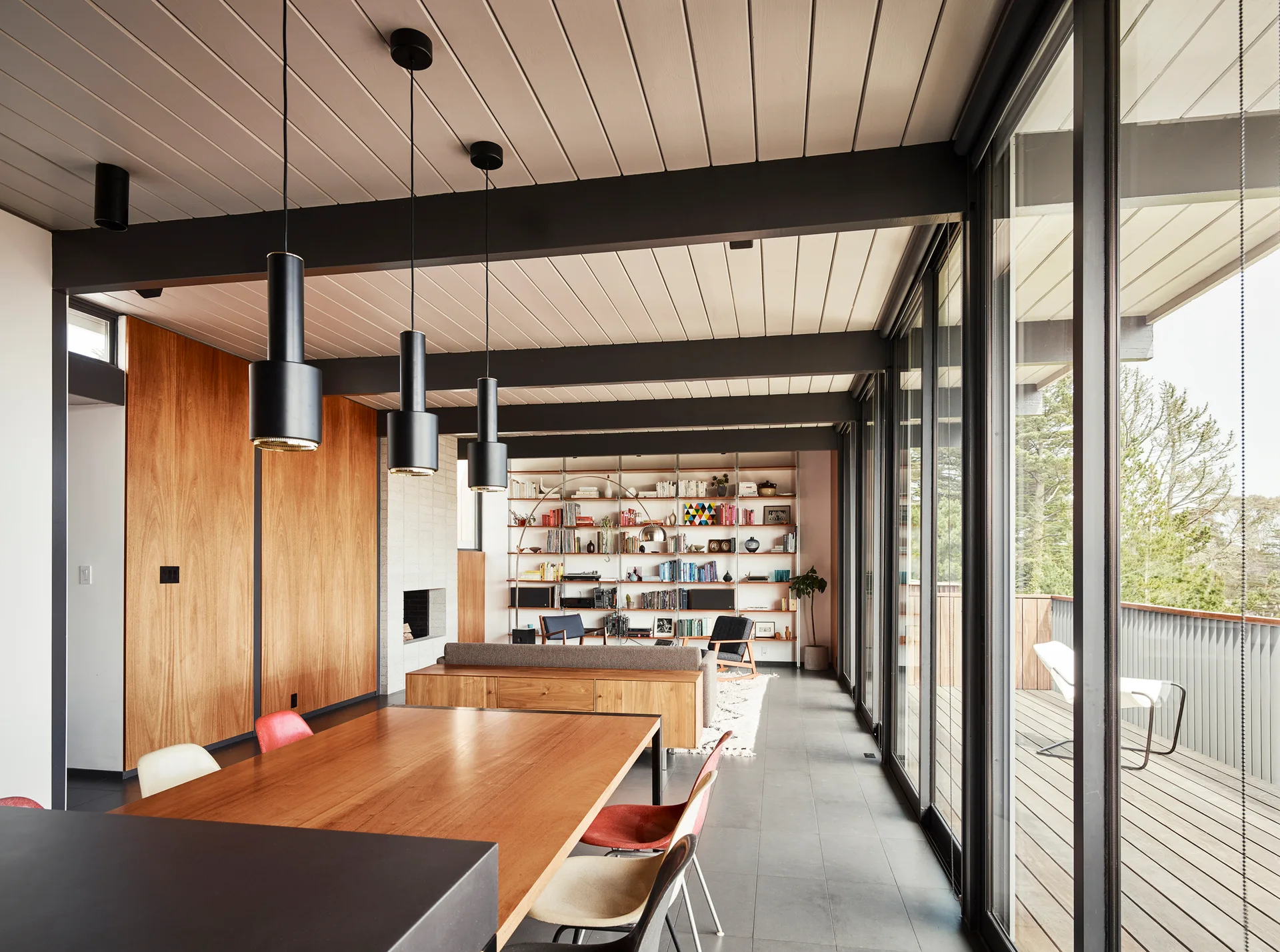 A mid-century modern open living space featuring a wood dining table under three pendant lights, exposed ceiling beams, and sliding glass balcony doors.