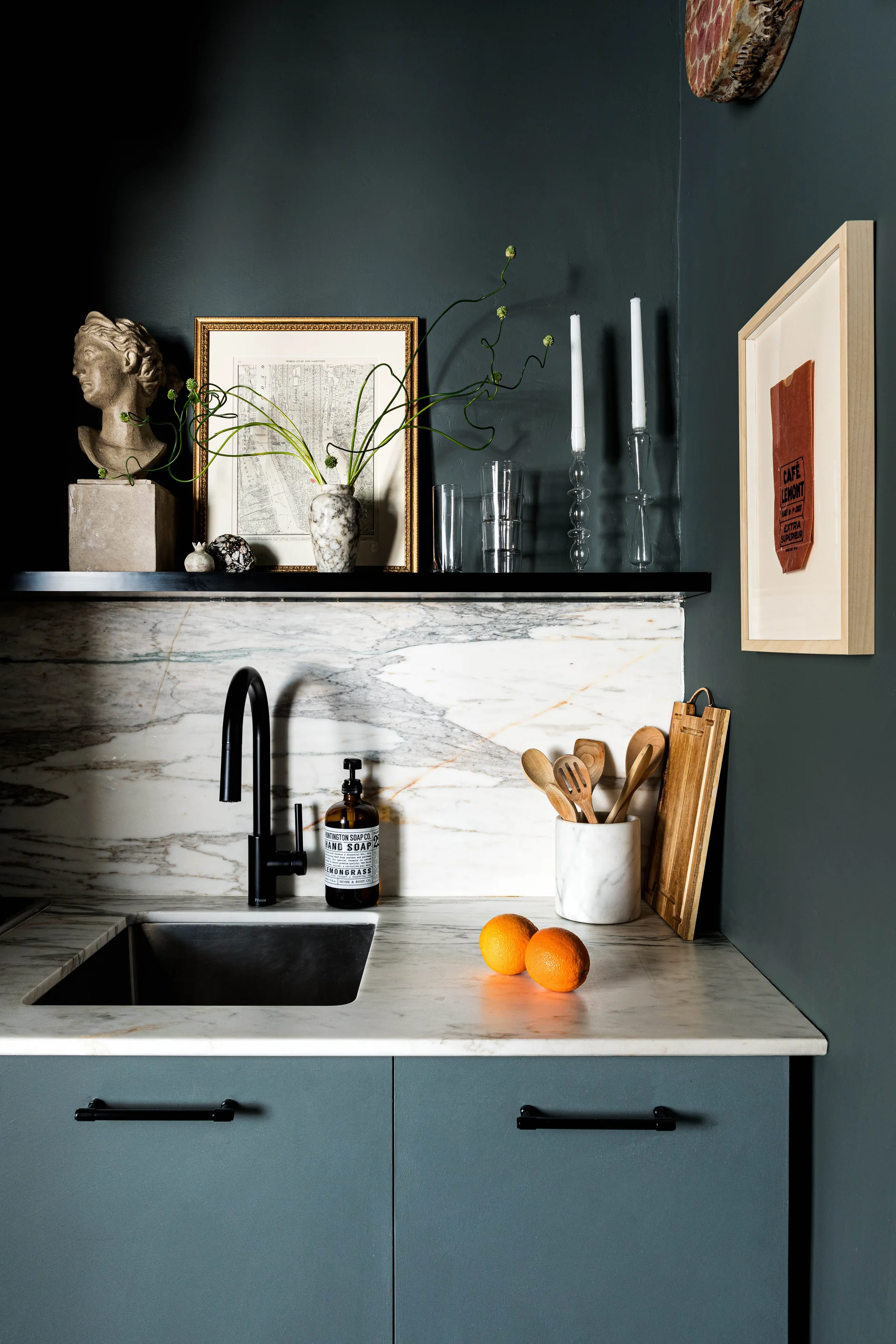 A kitchen sink area featuring a white veined marble backsplash, a black faucet, dark blue-grey cabinetry, and a styled black floating shelf.