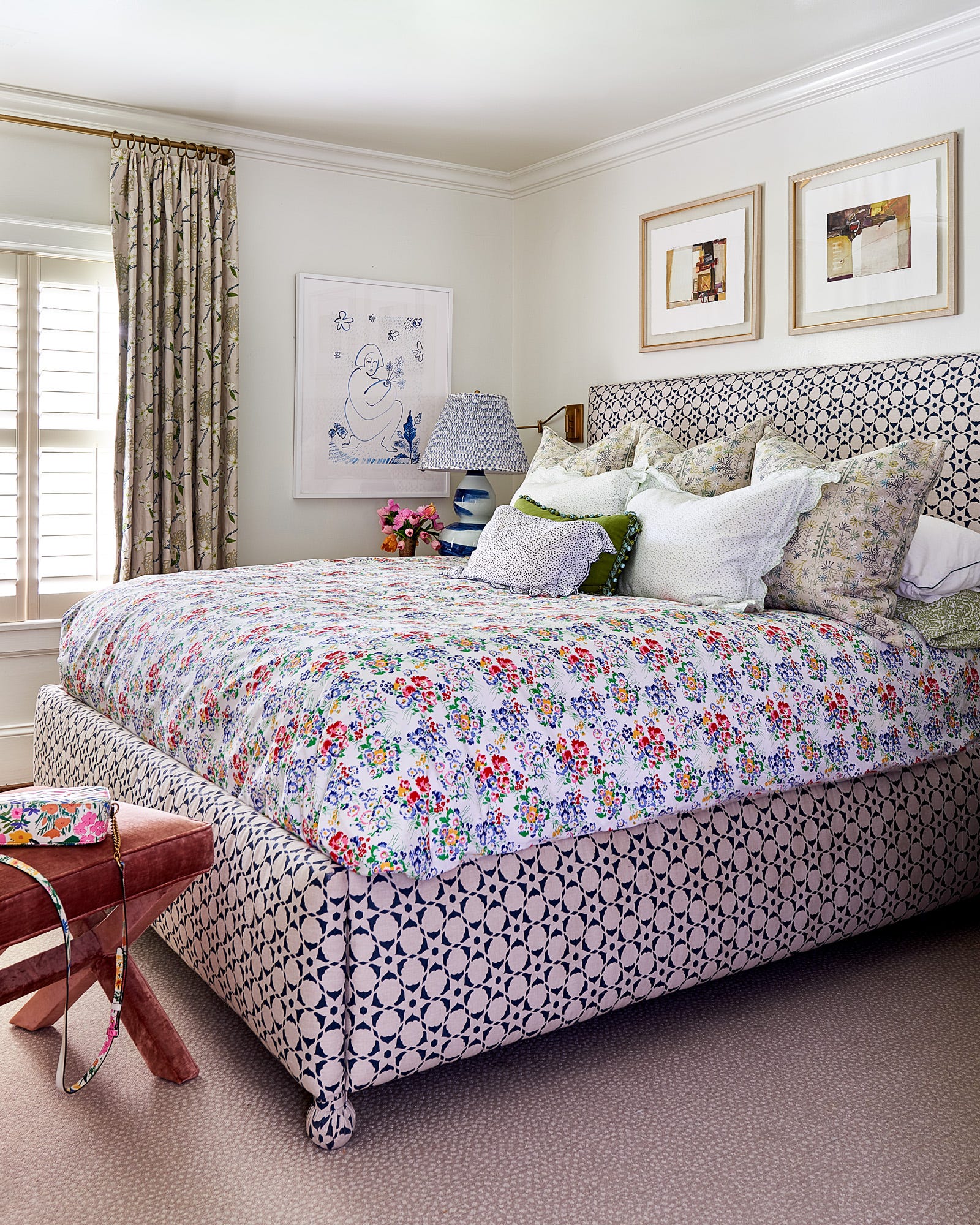 An upholstered bed with a blue geometric pattern and floral bedding sits next to a window with plantation shutters and patterned curtains.
