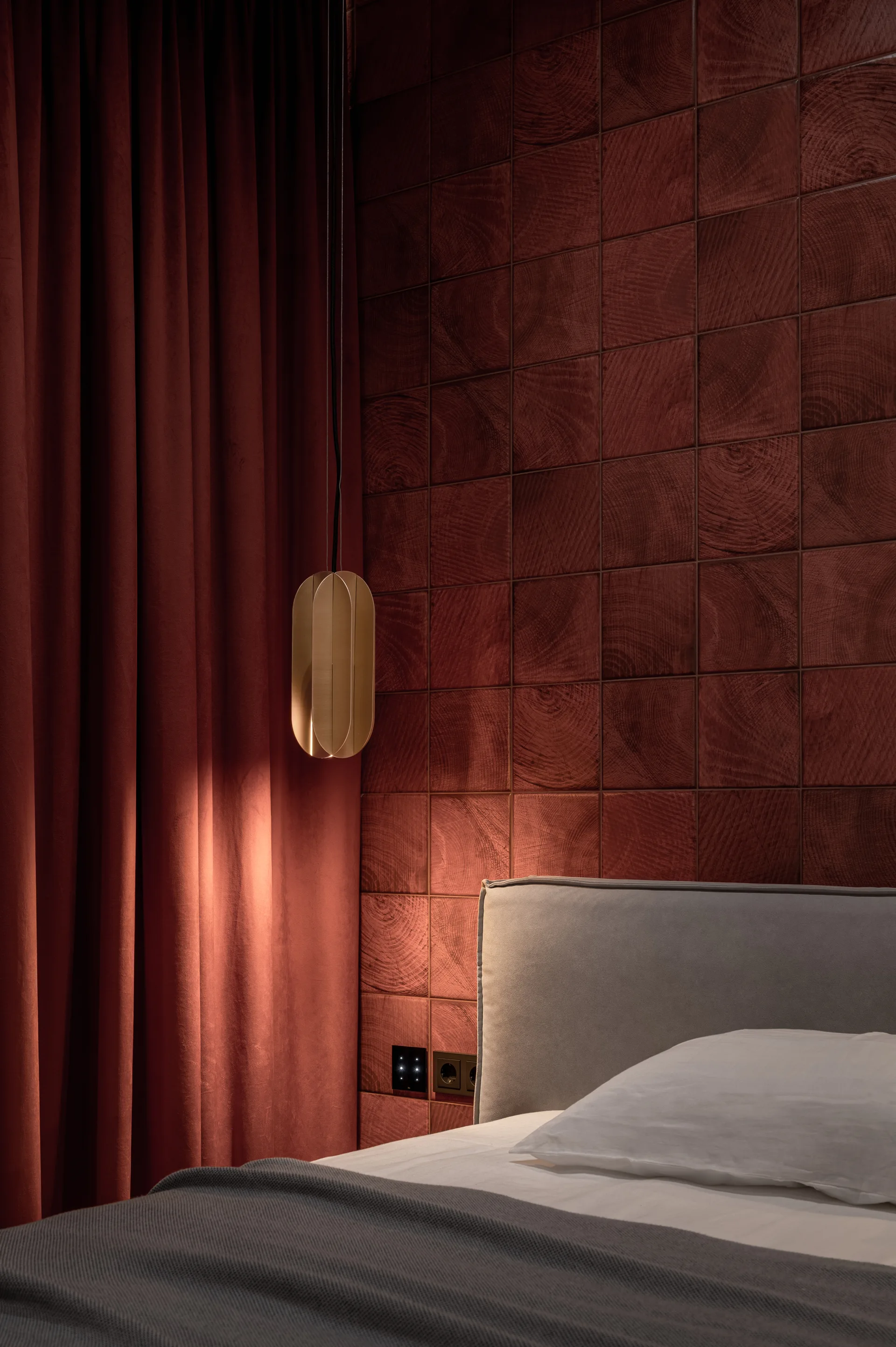 A bedroom features a deep red wooden tile accent wall, a gray upholstered headboard, matching red drapes, and a brass capsule pendant light.