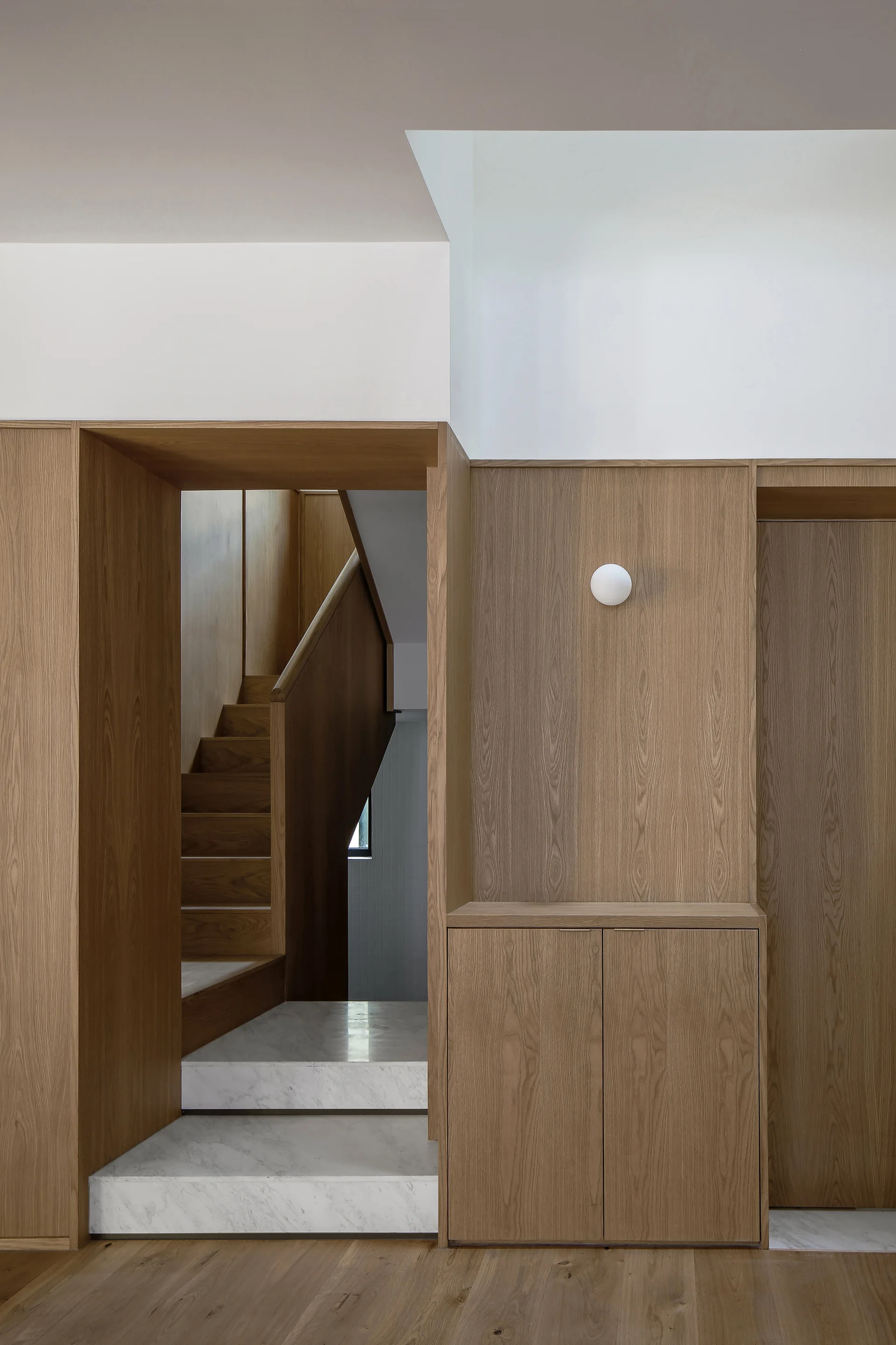 A modern hallway featuring warm wood paneling, a flush cabinet, a round white wall sconce, and white marble steps leading to a wooden staircase.