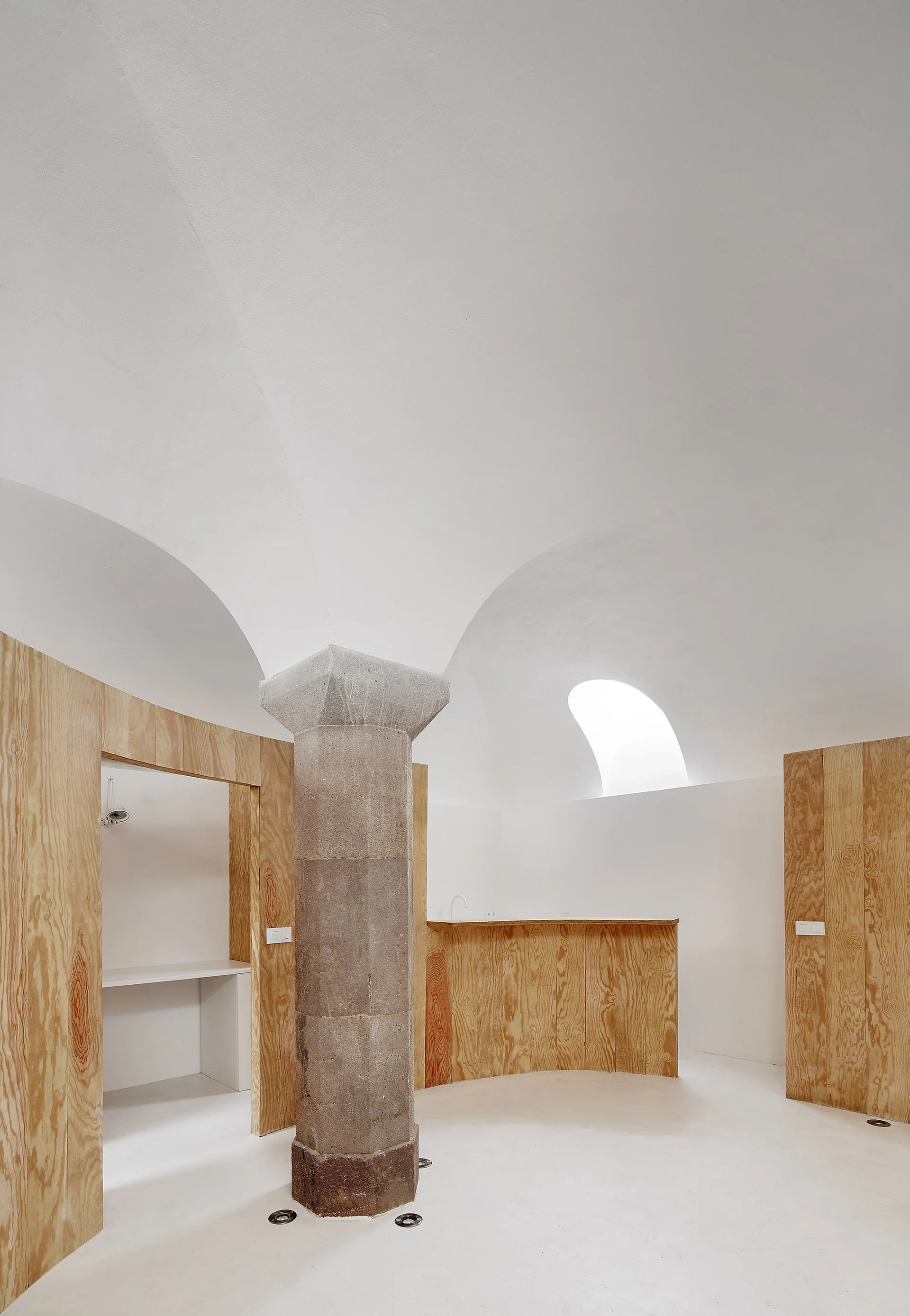 A minimalist room featuring an octagonal stone column under a white vaulted ceiling with light-toned plywood wall panels.