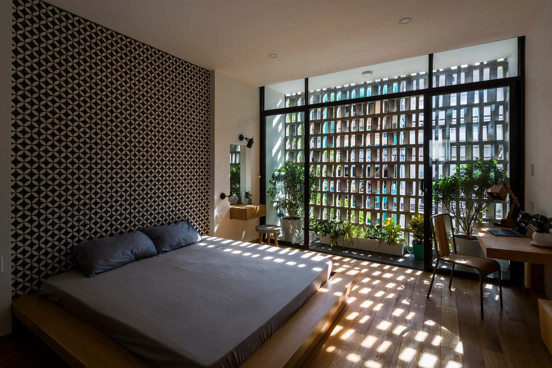 A minimalist bedroom features a low wooden platform bed, a geometric patterned accent wall, and glass sliding doors leading to a breeze block screen.