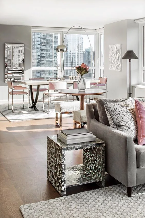 An open-concept apartment living room features a grey sofa and a dining table with pink chairs situated next to a large window.