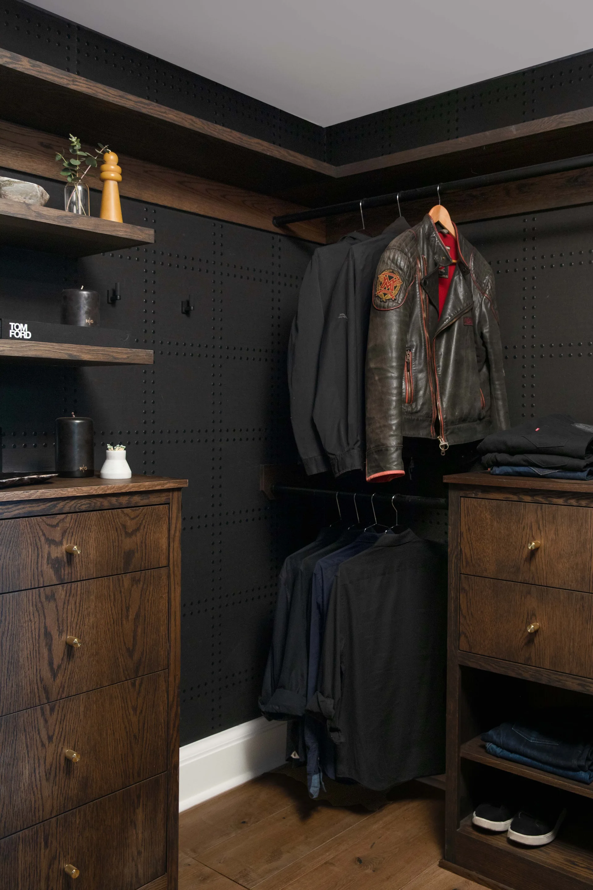 A walk-in closet featuring matte black pegboard walls, dark stained wood drawers with brass knobs, and black metal clothing racks.